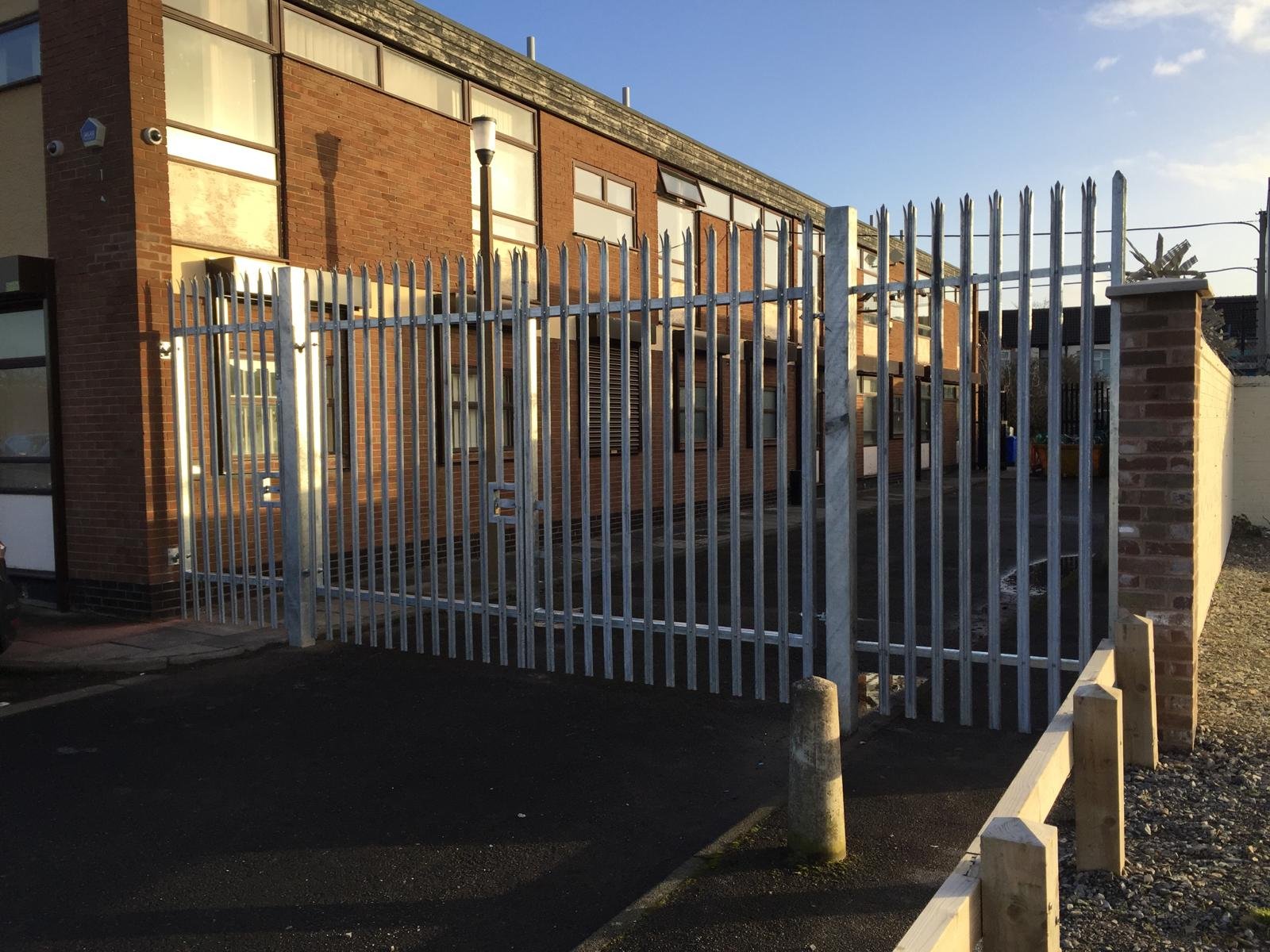 A metal fence with pointed tips surrounding a brick building. The building has large windows and a small doorway. There is a lamppost and some construction elements in the foreground.