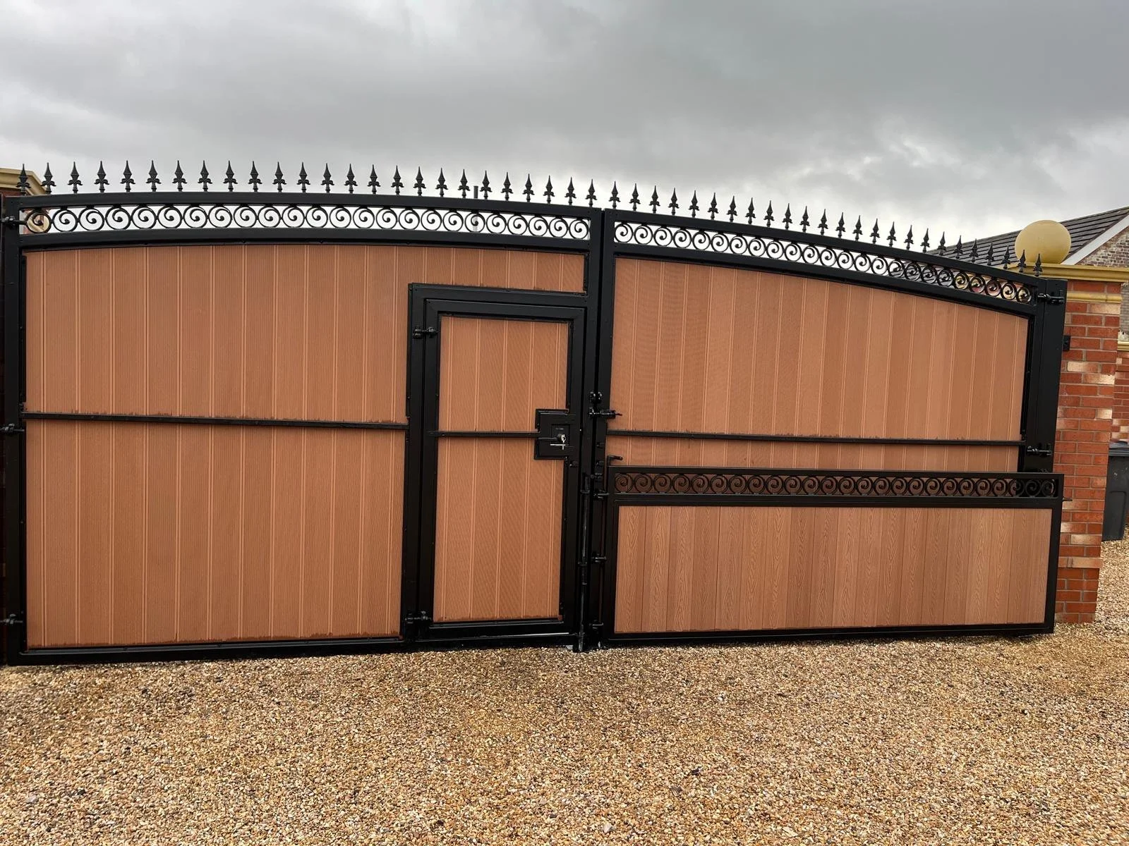 A large gate with a black metal frame and decorative ironwork, featuring a wooden panel design in a residential area with a gravel driveway and a brick wall on the right.