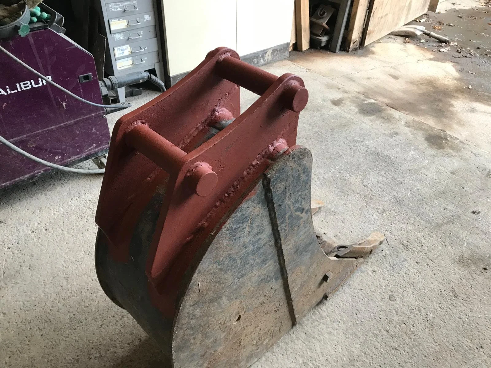 A close-up of a red hydraulic excavator bucket attachment resting on a concrete floor in a workshop.