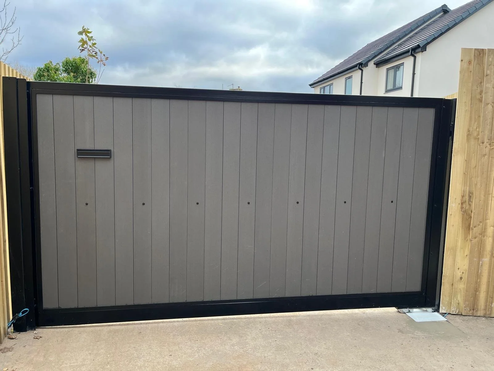 A modern gray sliding gate with black framing, installed in front of a residential property with white walls and a tilled roof house in the background, under a cloudy sky.