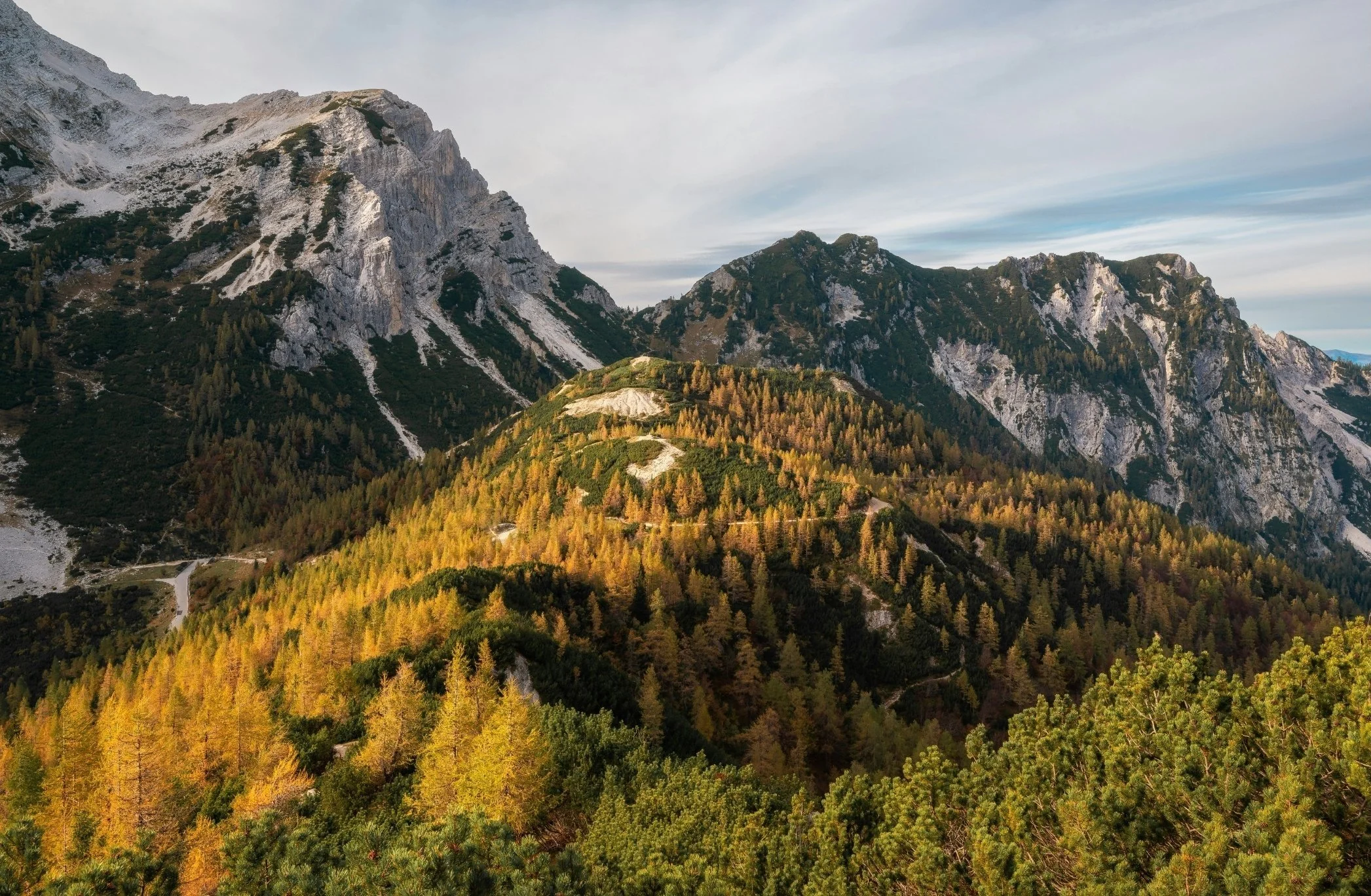Landscape of mountains with rocky peaks, forested slopes, and a winding trail, under a partly cloudy sky.