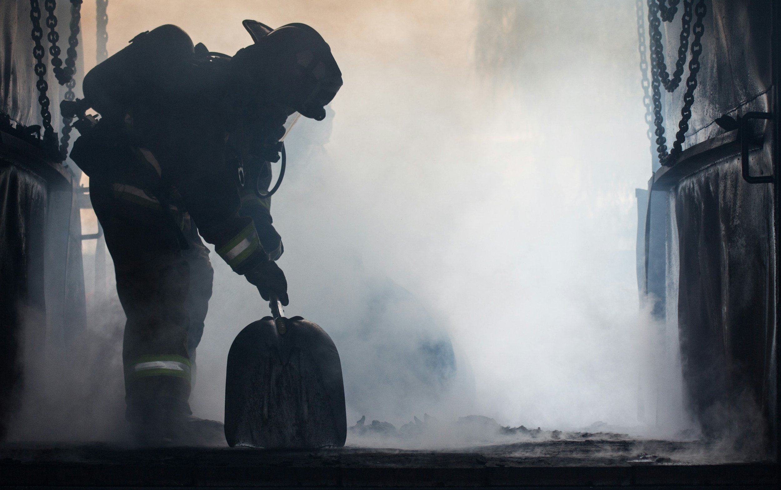Firefighter in full gear spraying water during a fire emergency, surrounded by smoke and chains hanging from the building ceiling.