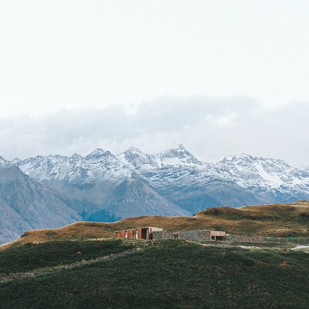 Modern house on a grassy hillside with snow-capped mountains in the background.