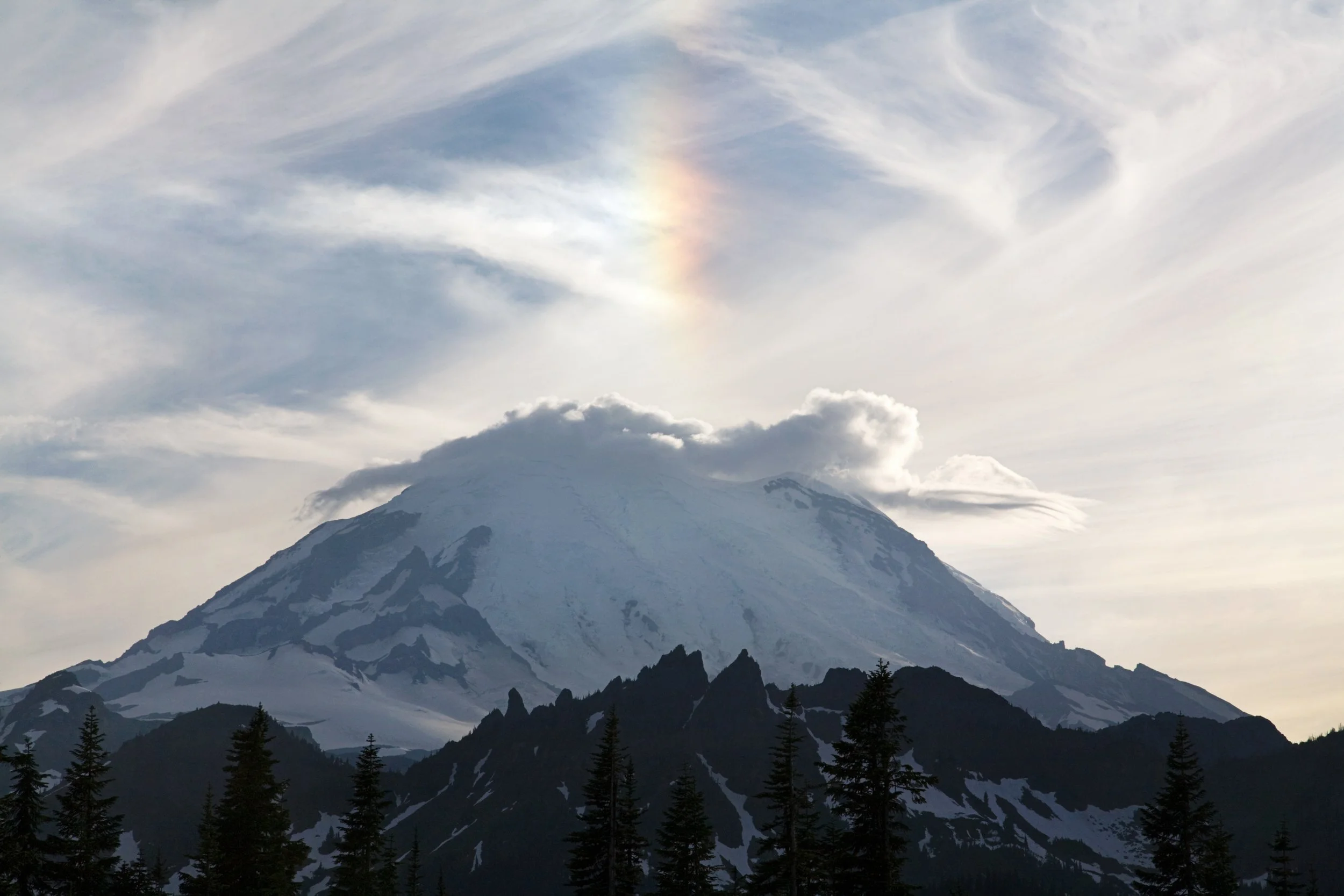 Snow-capped mountain with forested foreground and a rainbow in the cloudy sky.