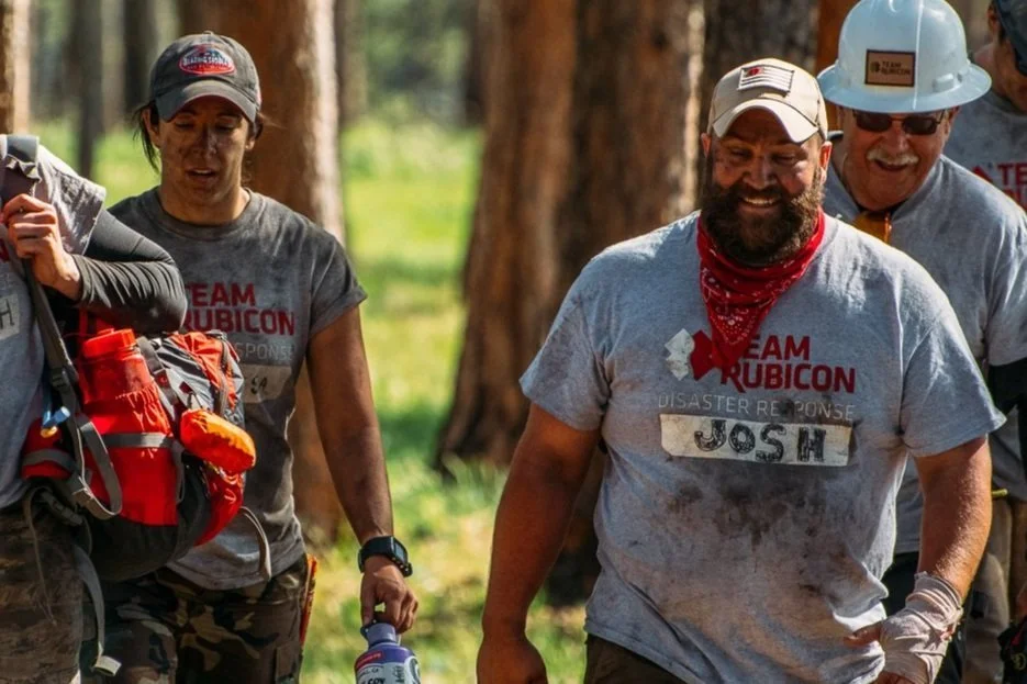 Group of smiling people participating in a team-building outdoor activity in a forest, wearing matching gray T-shirts with 'Team Rubicon' and 'Disaster Response' written on them, some with helmets and gear.