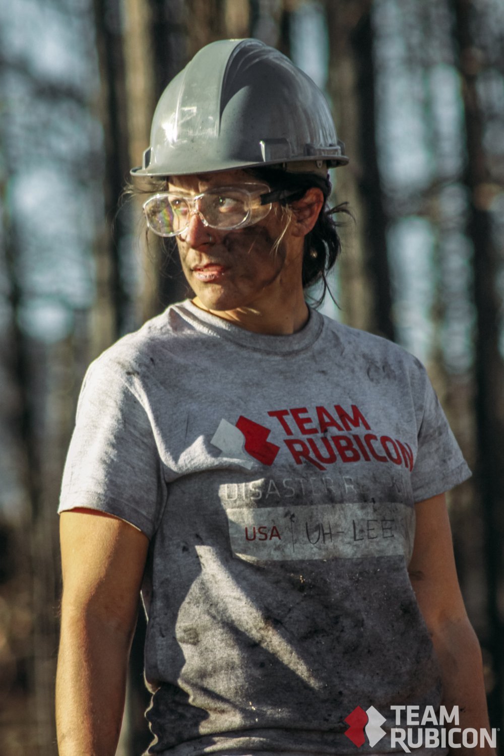 Female worker wearing a gray helmet, clear safety glasses, and a gray t-shirt with 'TEAM RUBICON' logo in red, standing in a forested area.