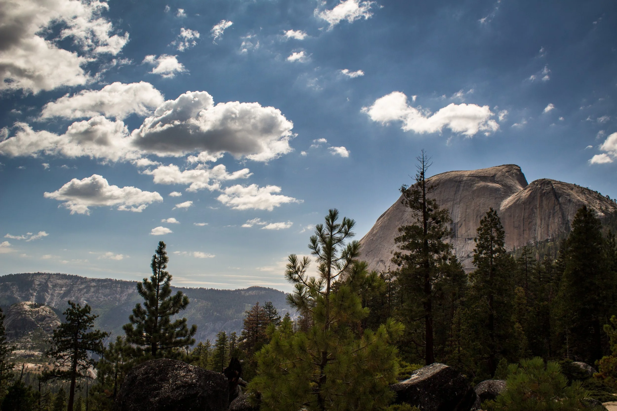 A mountainous landscape with large granite domes, tall trees, and a partly cloudy sky.