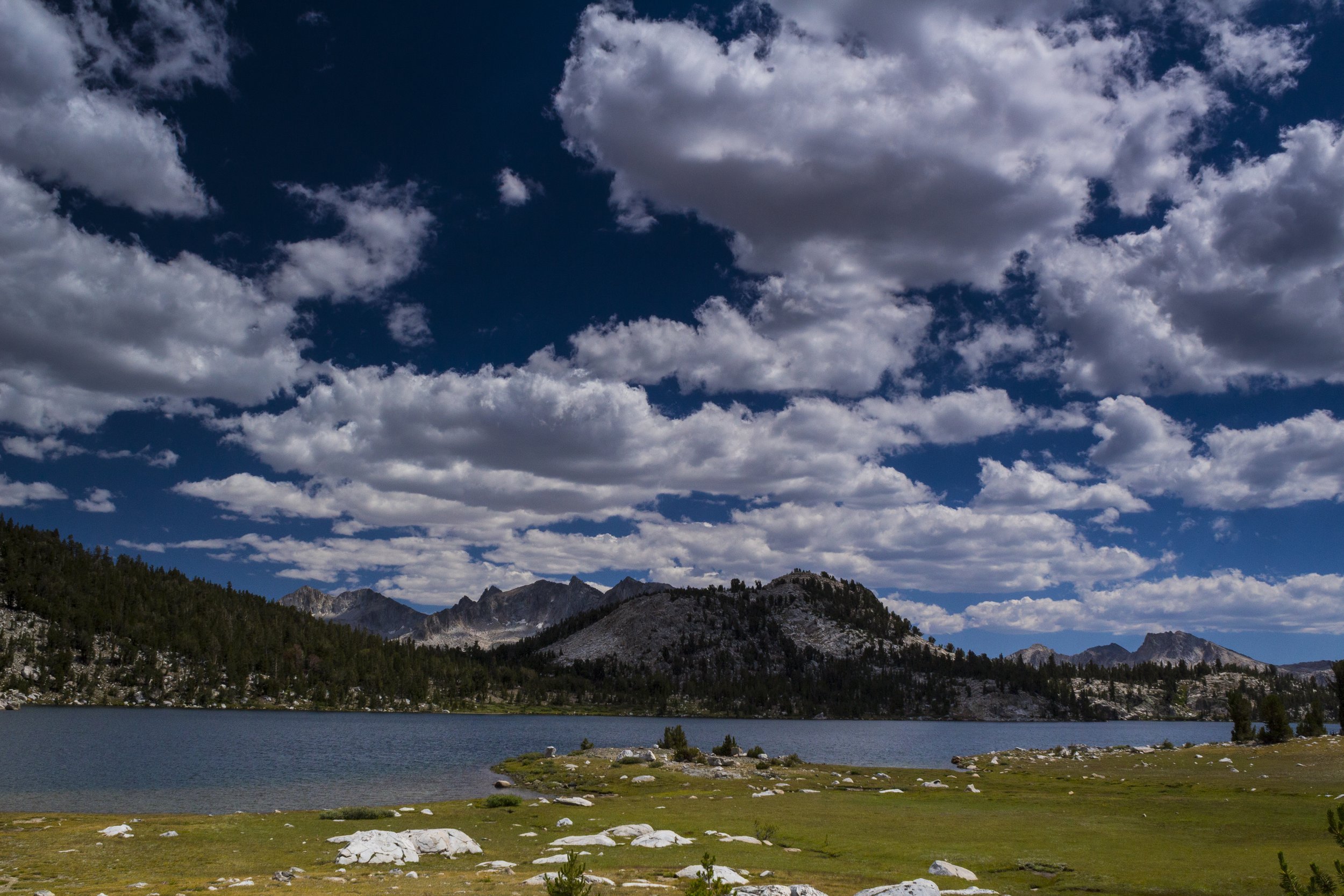 Scenic landscape of a mountain lake with a forested shoreline in the foreground, and rugged mountains under a partly cloudy sky in the background.