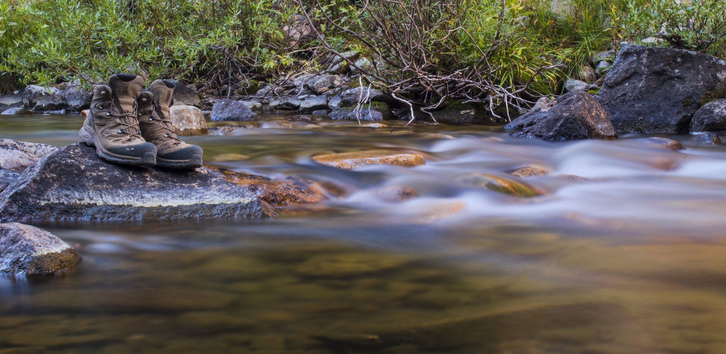 A pair of hiking boots resting on a rock in a flowing mountain stream, with green foliage and rocks in the background.