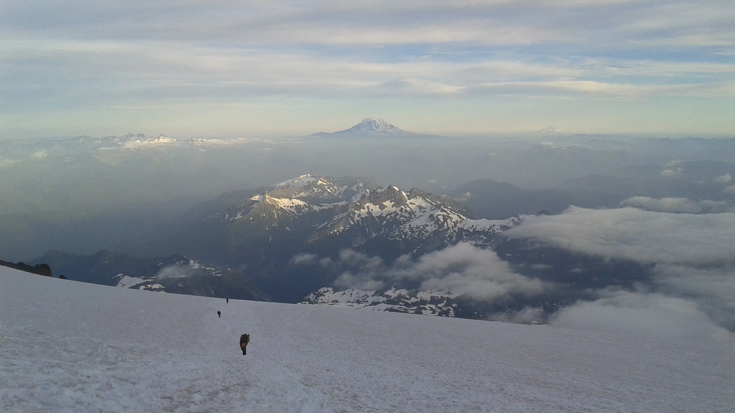 Snow-covered mountain slope with hikers walking, mountain peaks in the background, clouds, and a distant volcano