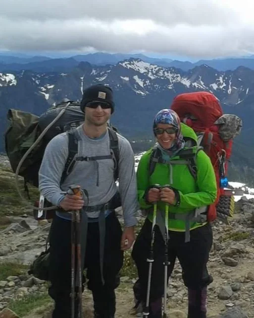 Dr. Alisa Bartel and brother in outdoor gear standing on a rocky mountain trail with snow-capped peaks and cloudy sky in the background.