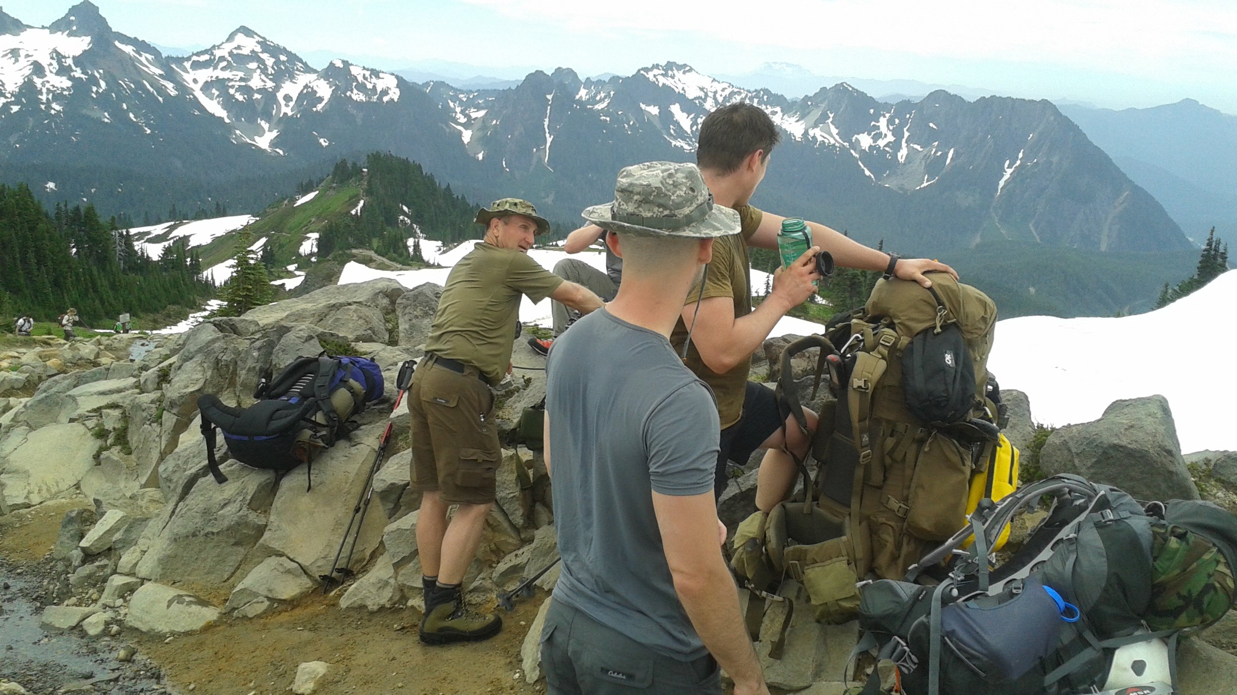 Three men on a mountain ridge with military backpacks, rocks, and hiking gear, overlooking snow-capped mountains and green forested slopes in the distance.