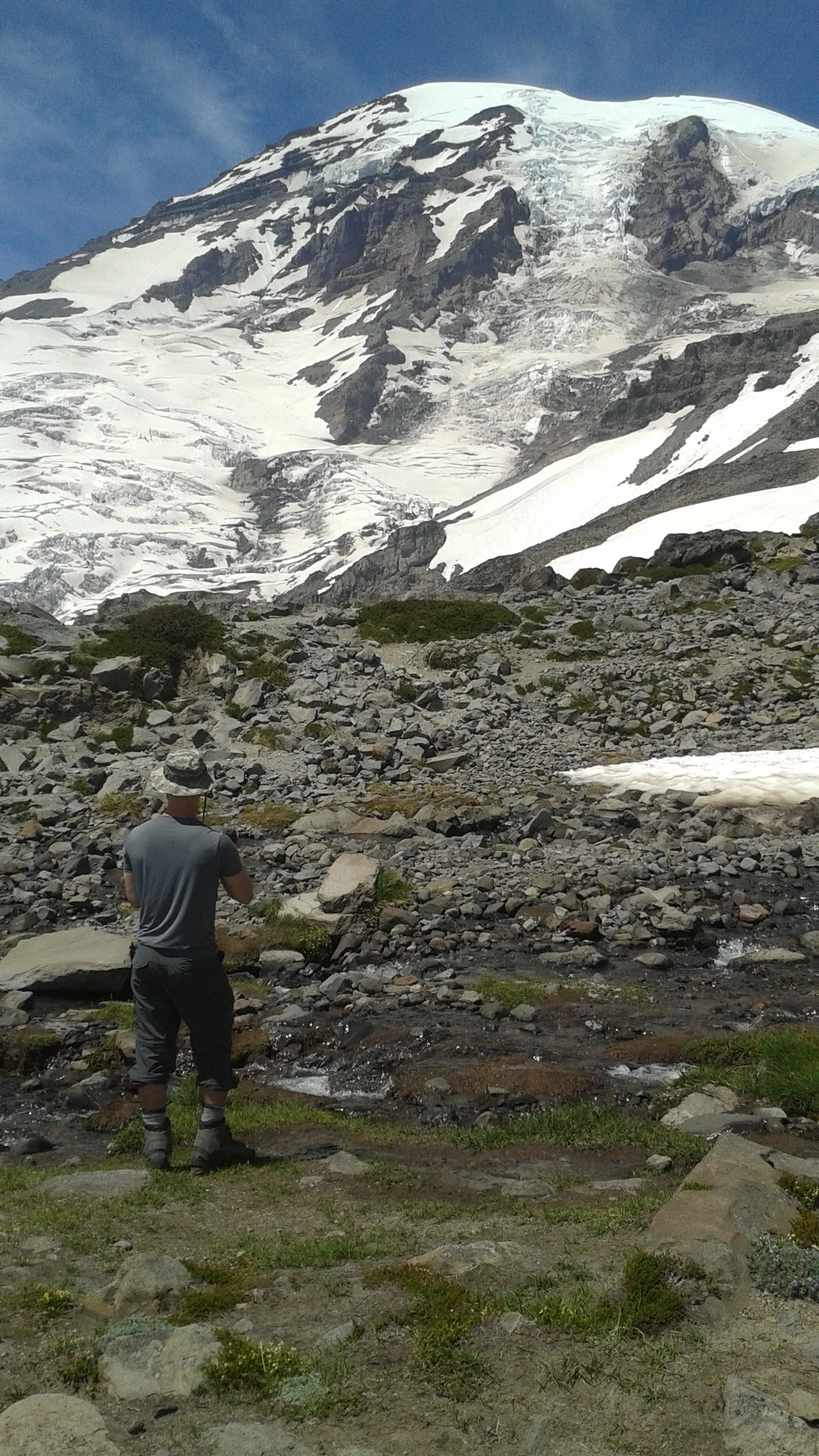 Person standing on rocky terrain near snow-capped mountain with glacier in the background of Mt. Rainier.