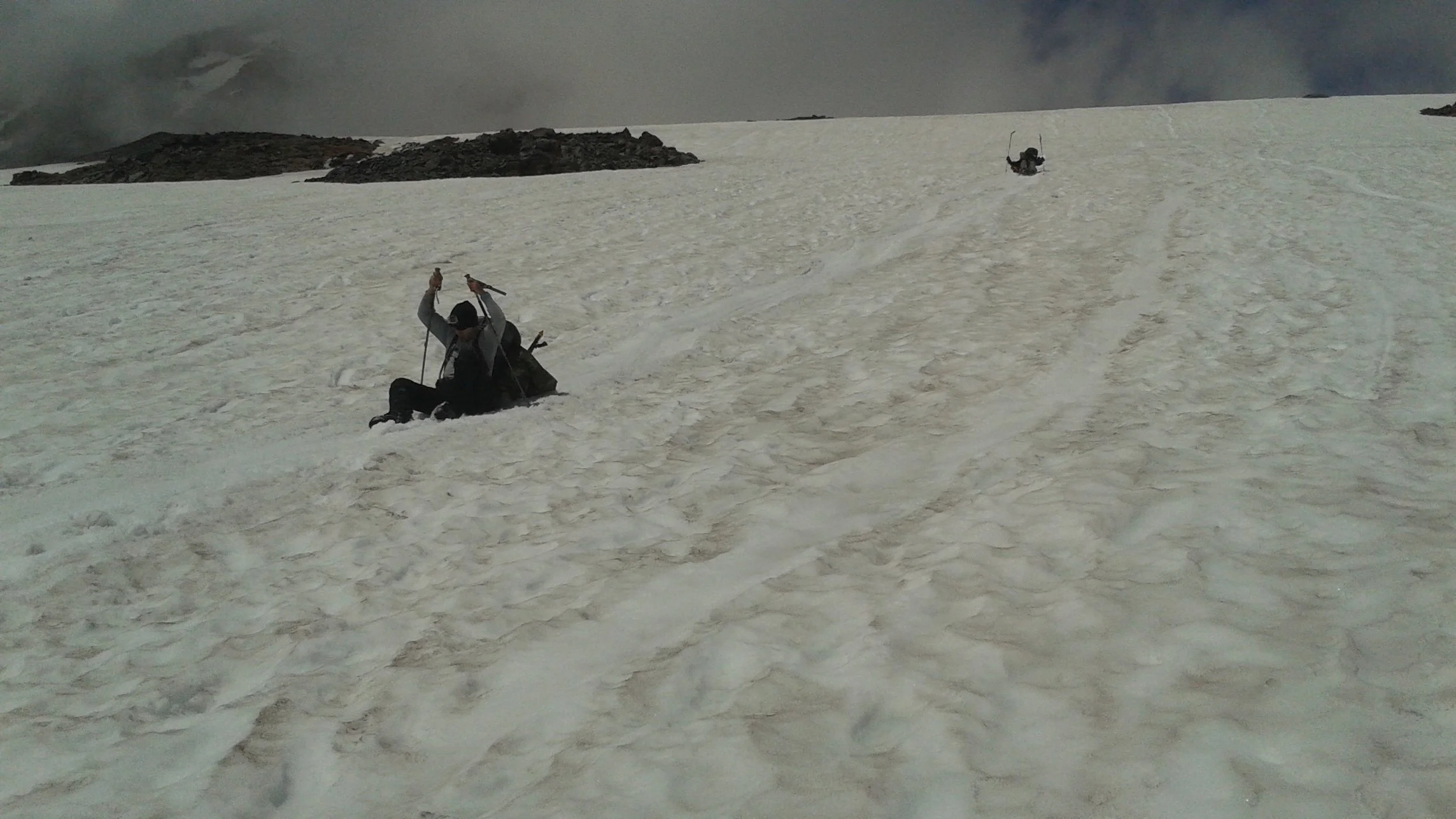 A person sliding down on snow in a mountainous, glacial landscape holding trekking poles, with another person in the background near climbing gear on a snowy slope of Mt. Rainier.