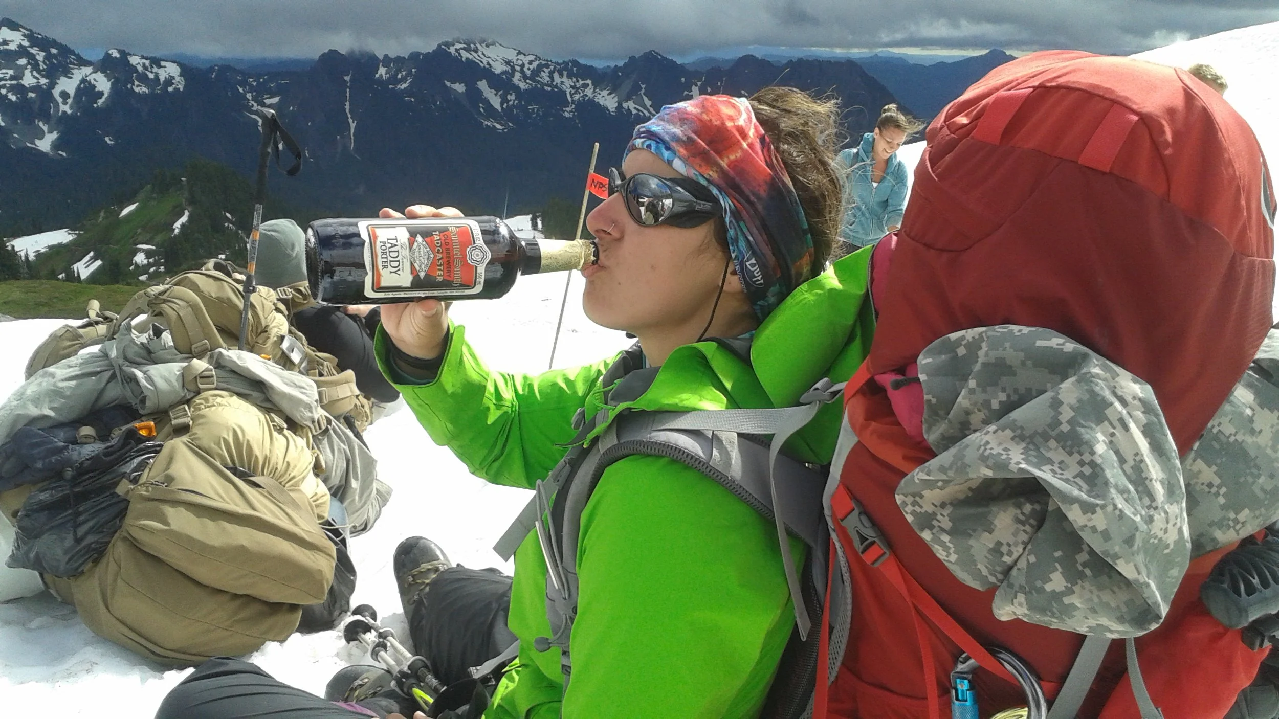 Dr. Alisa Bartel in a bright green jacket, wearing sunglasses and a colorful headband, sitting on snow in a mountain landscape, drinking from a bottle of celebratory beer amidst other hikers and gear.