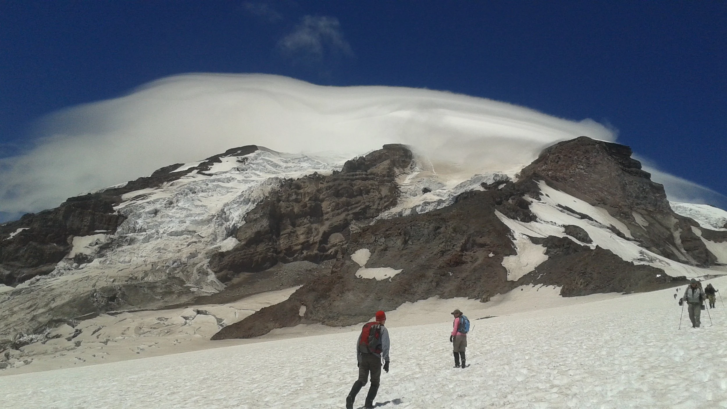 Group of hikers walking on snow-covered terrain near a mountain with a glacier and a large snow cloud above Mt. Rainier.
