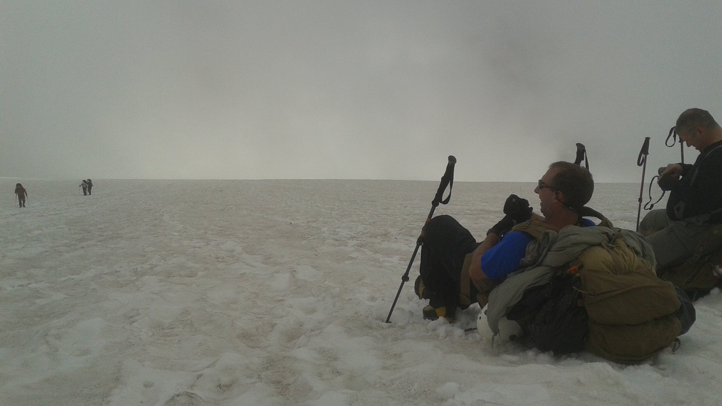 Group of hikers resting on a snow-covered landscape with backpacks, some sitting and some standing, under cloudy sky on Mt. Rainier.