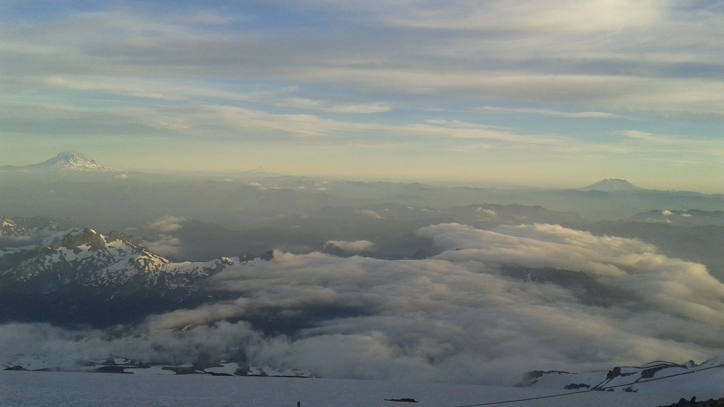 Aerial view of snow-capped mountains with clouds and distant volcanoes on the horizon under a partly cloudy sky.