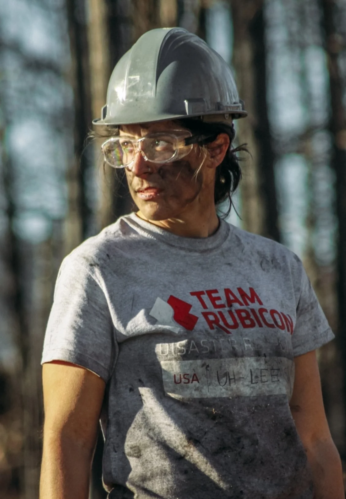 Dr. Alisa Bartel, psychologist in Colorado Springs, wearing a grey T-shirt with 'Team Rubicon' logo, safety goggles, and a hard hat standing outdoors in a forest.