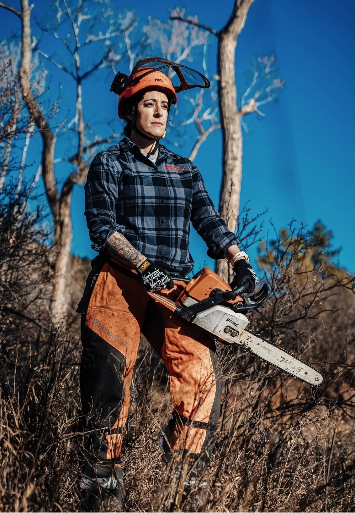 Dr. Alisa Bartel, psychologist in Colorado Springs, in outdoor gear holding a chainsaw in a forested area with burnt trees and dry grass, under a clear blue sky.