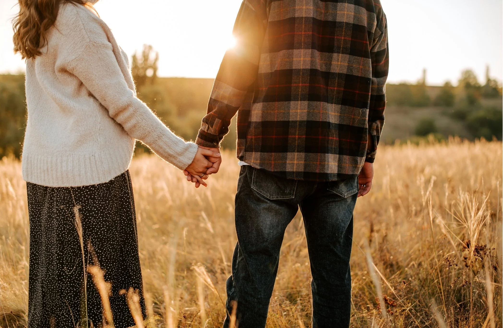 Two people holding hands in a field during sunset, sunlight shining between them.