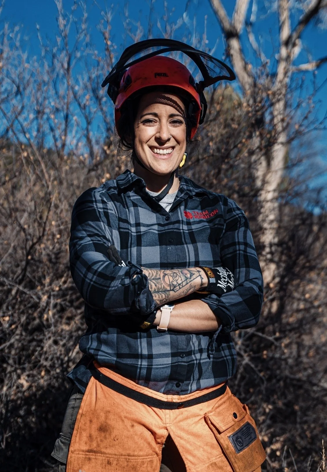 Dr. Alisa Bartel, psychologist in Colorado Springs, smiling, wearing a red helmet with a face shield, a plaid shirt, and orange work pants, standing outdoors with leafless trees in the background.