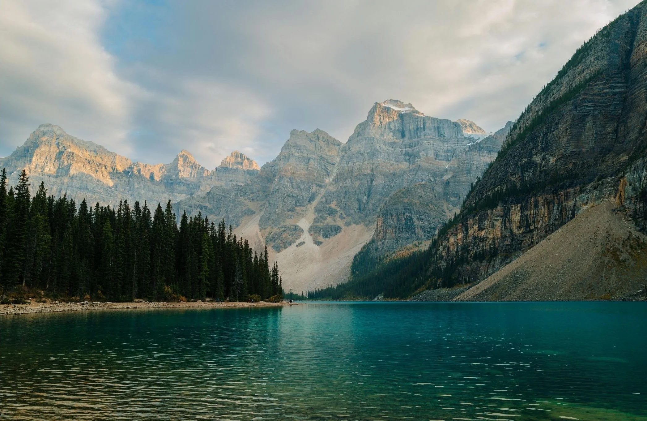 A serene mountain landscape with a calm lake in the foreground, dense evergreen trees along the shoreline, and towering snowy mountain peaks under partly cloudy skies.
