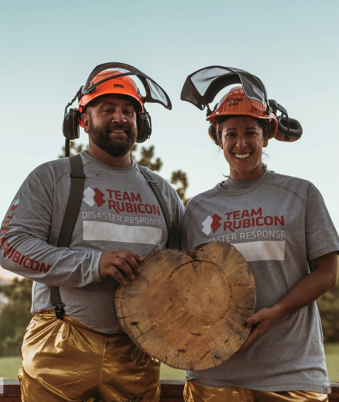 Two people wearing orange helmets and gray shirts that say 'Team Rubicon Disaster Response' are holding a large piece of wood with a tree ring pattern, smiling outdoors during sunset.