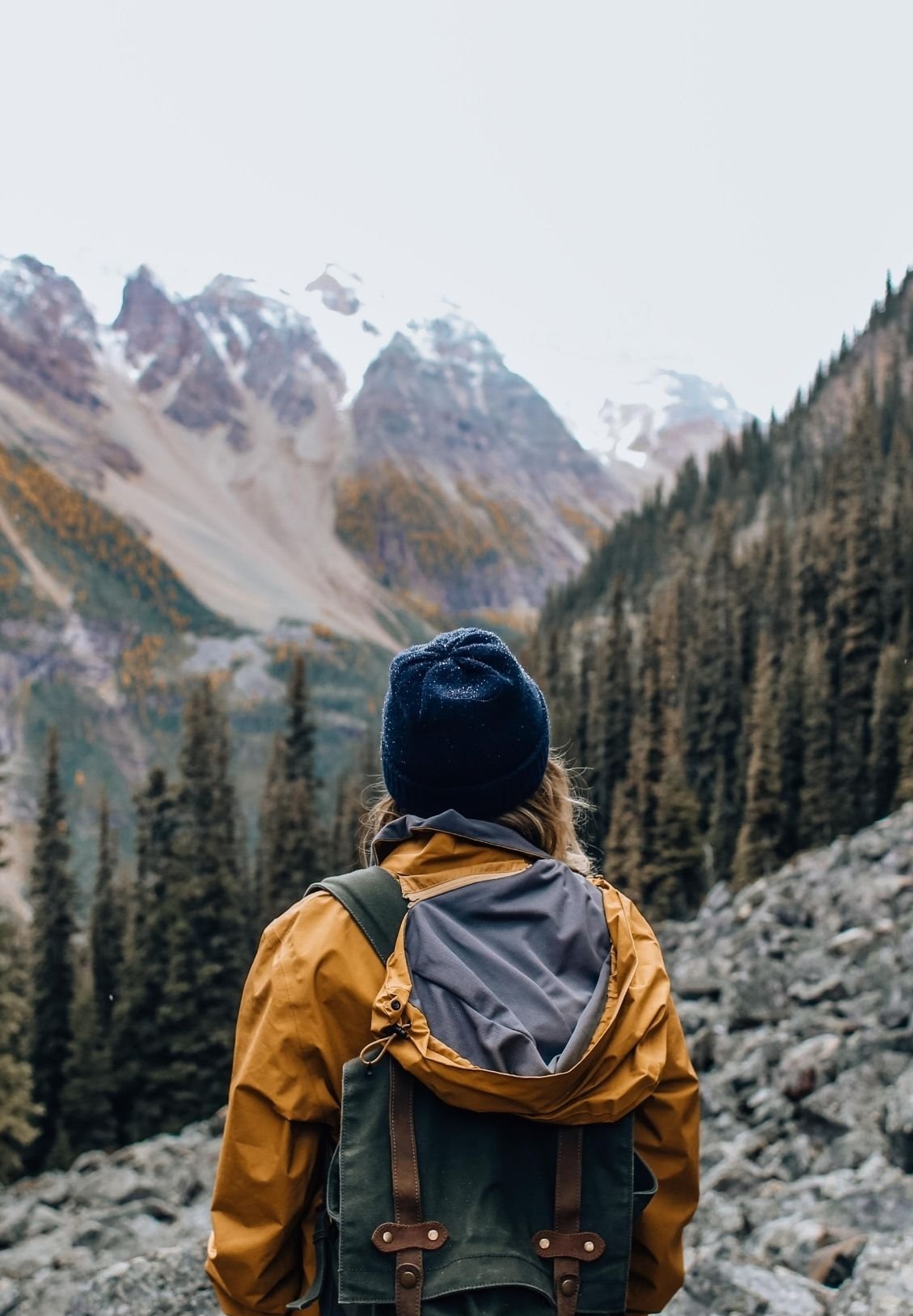 A hiker wearing a black beanie, yellow jacket, and backpack looking at snow-capped mountains and forested valley.