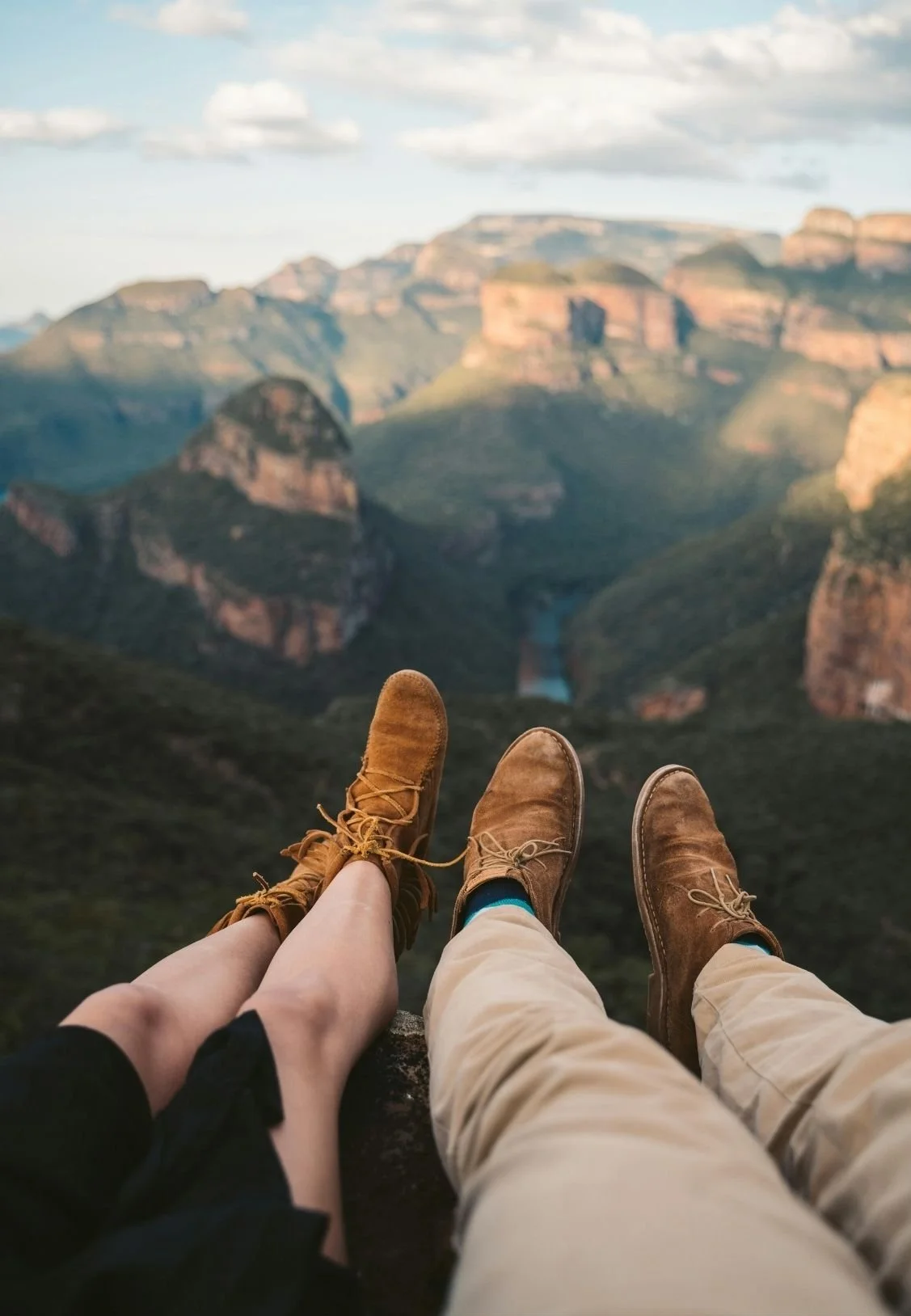 Two people wearing brown boots sitting on a ledge overlooking a canyon with green hills and a waterfall, under a partly cloudy sky.
