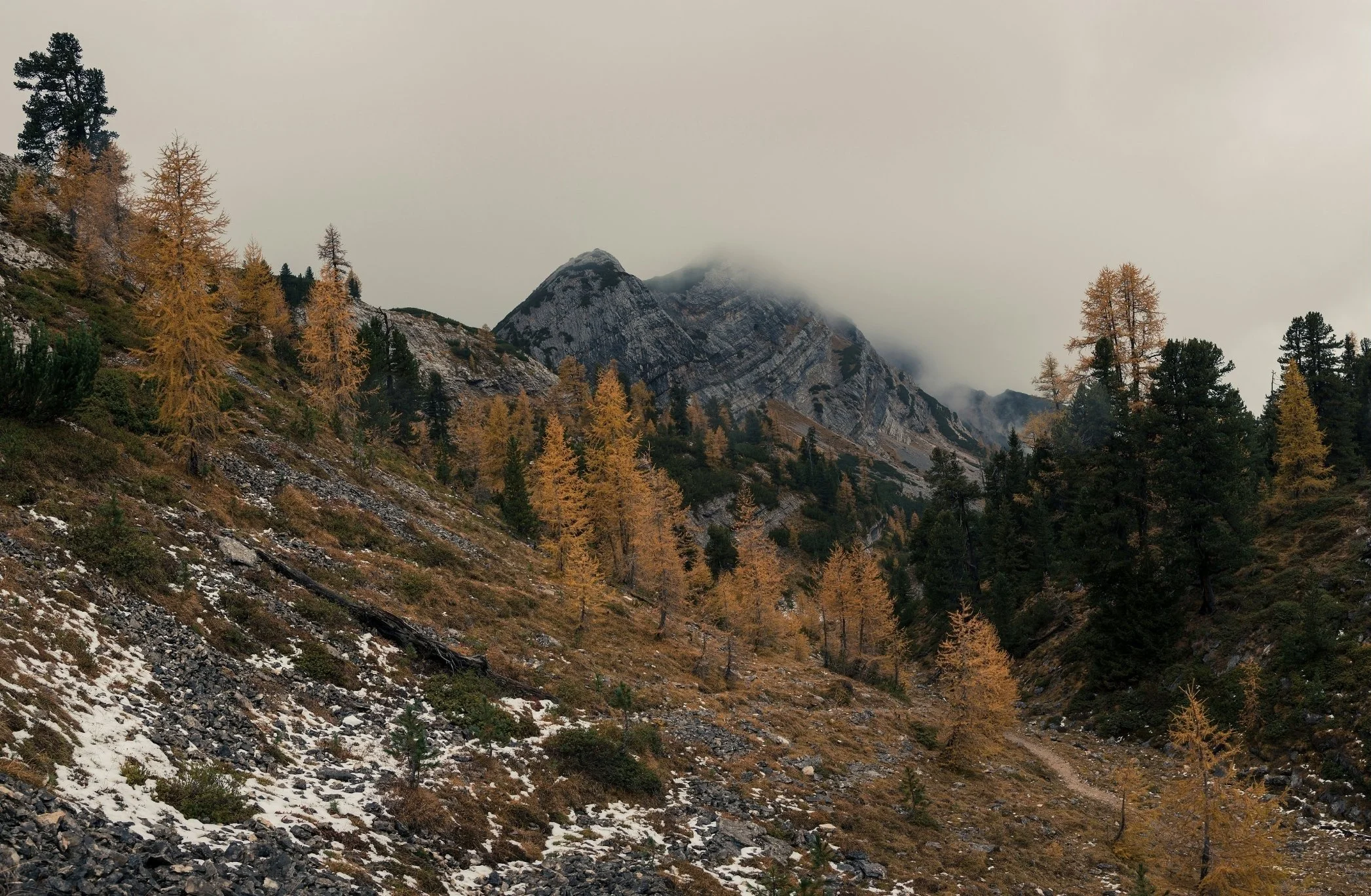 Mountain landscape with rocky peaks partially covered by fog, surrounded by evergreen and deciduous trees in autumn colors, some snow on the ground.