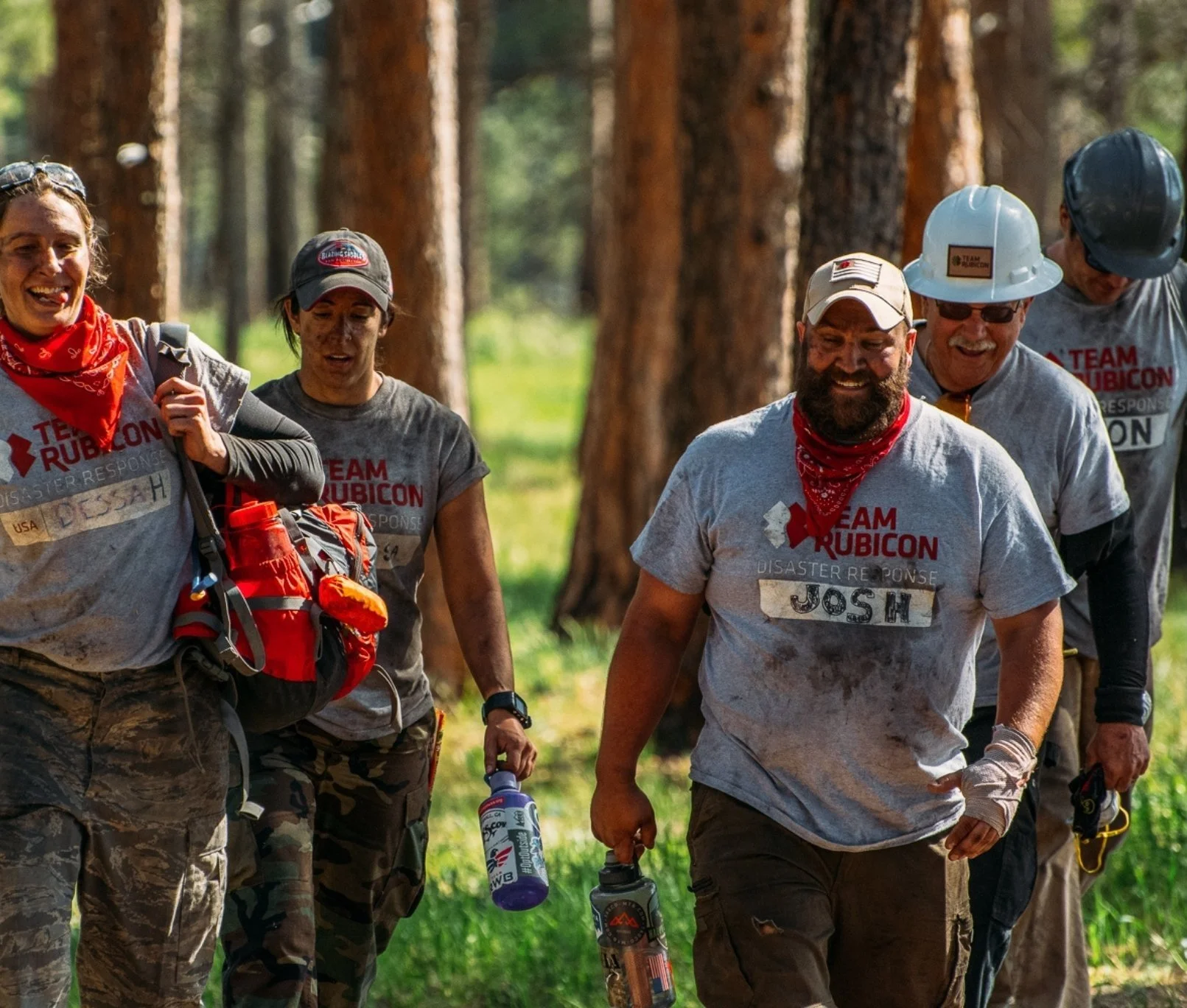 Group of people hiking in a forest, wearing gray 'Team Rubicon' shirts, some with helmets and carrying water bottles.