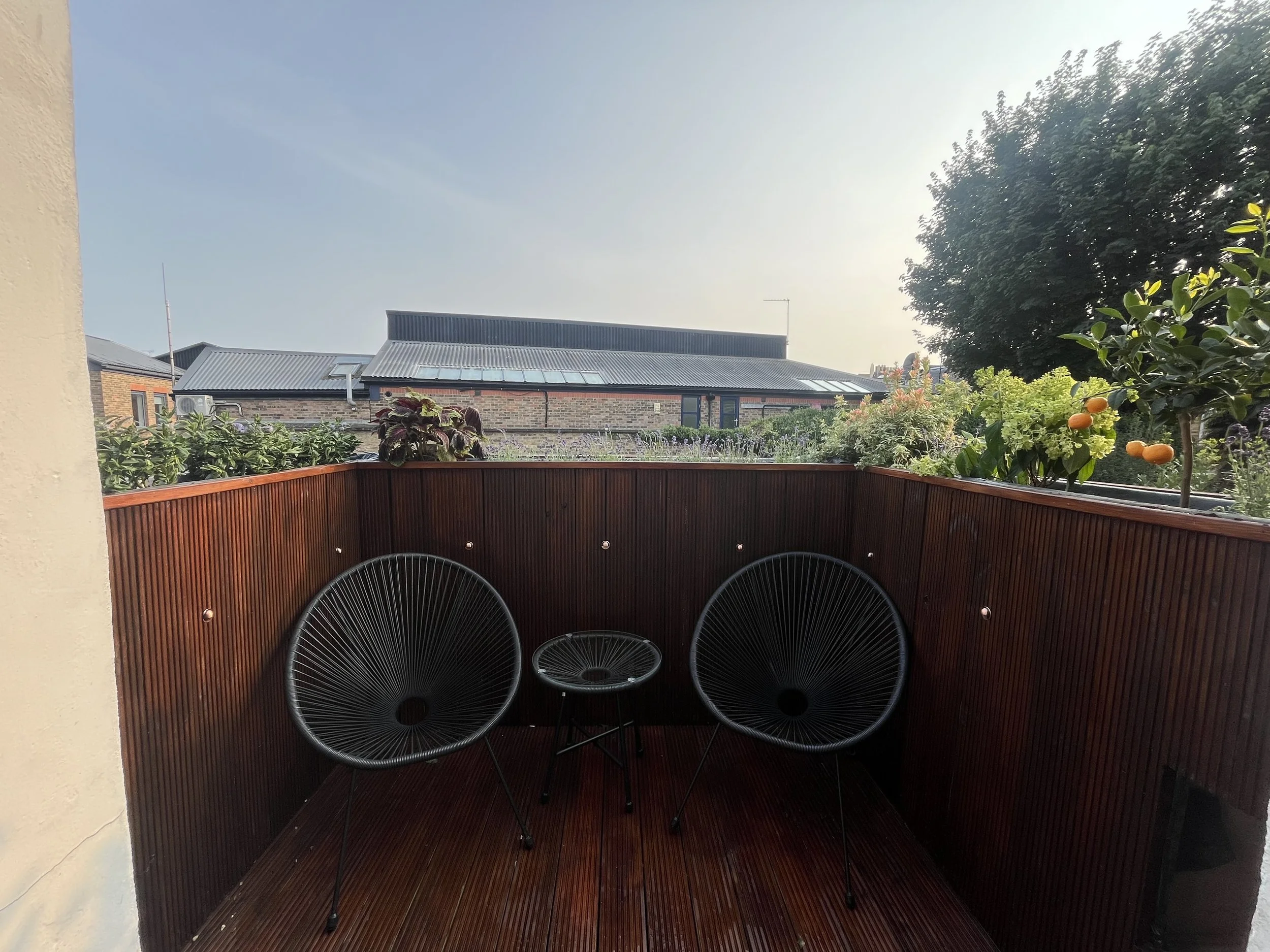 Balcony with redwood railing, black chairs, flower pots, garden with bushes, tree, and neighboring buildings in the background under a blue sky.