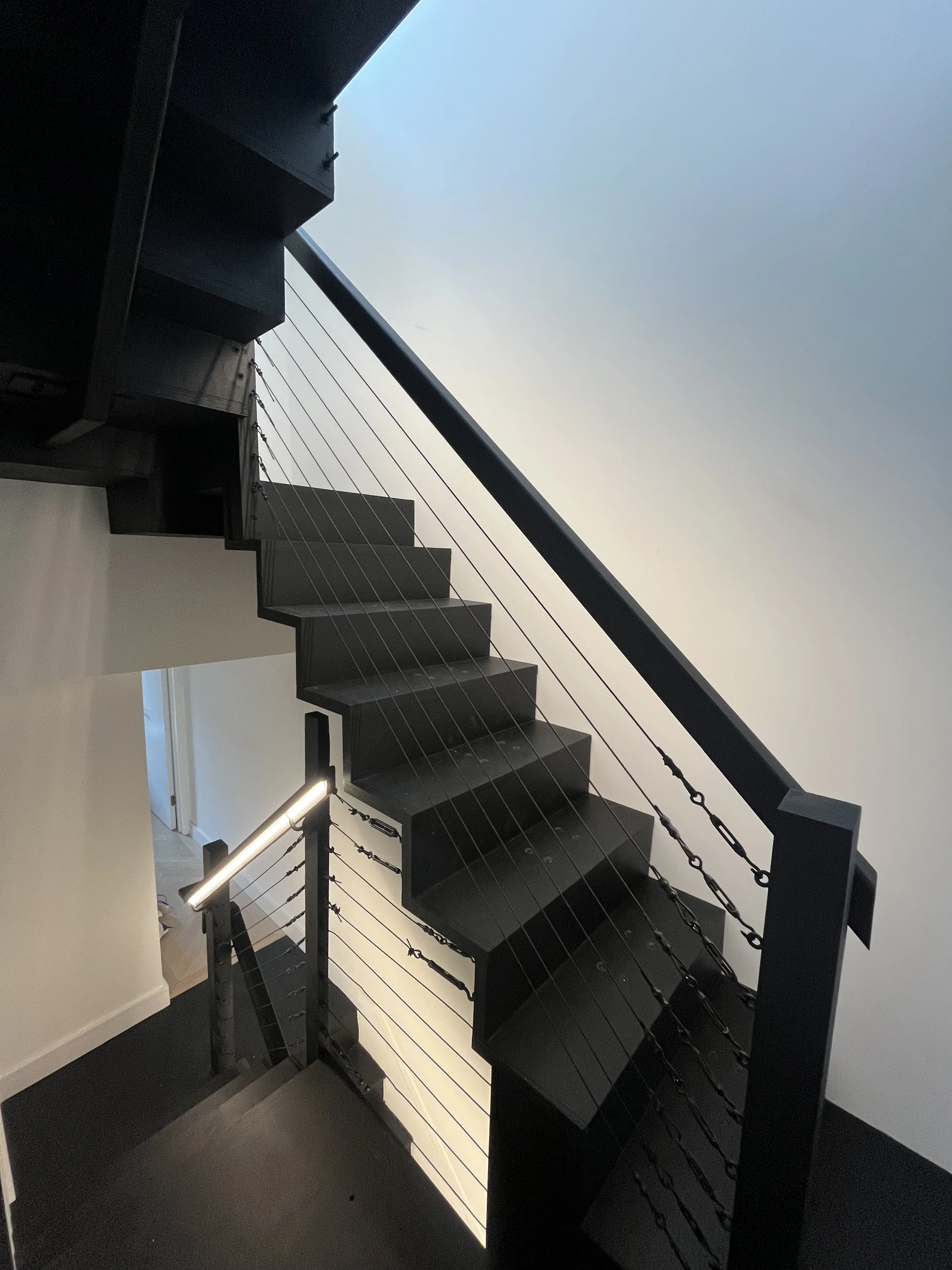 A modern black staircase with open risers, metal chains as railings, and a white wall with a door in the background.
