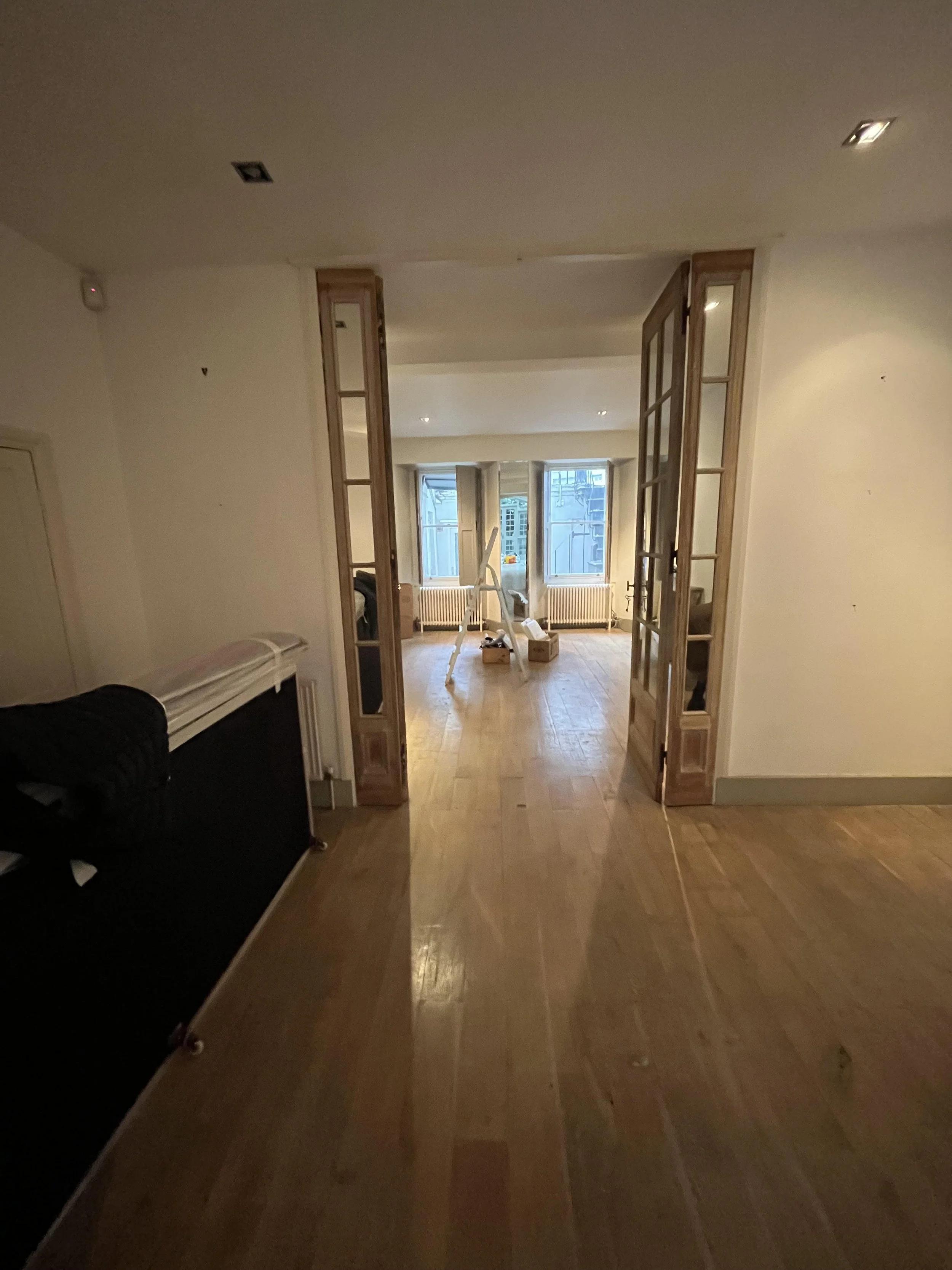 View of an interior room with wooden floors, leading to a bright space with large windows, some tools and materials are present, indicating ongoing renovation or decorating.