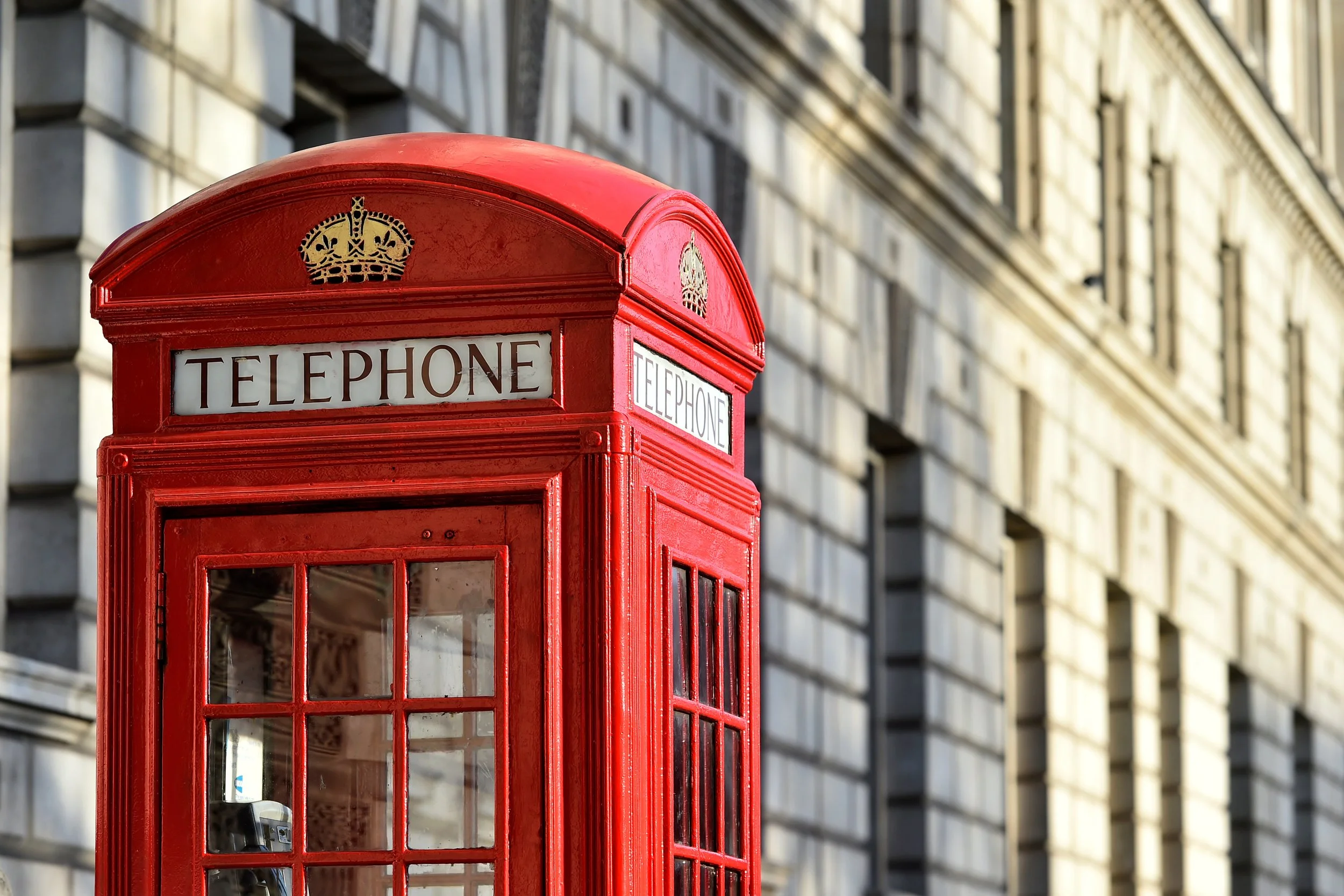 A classic red British telephone booth on a city street with a background of a large stone building with multiple windows.