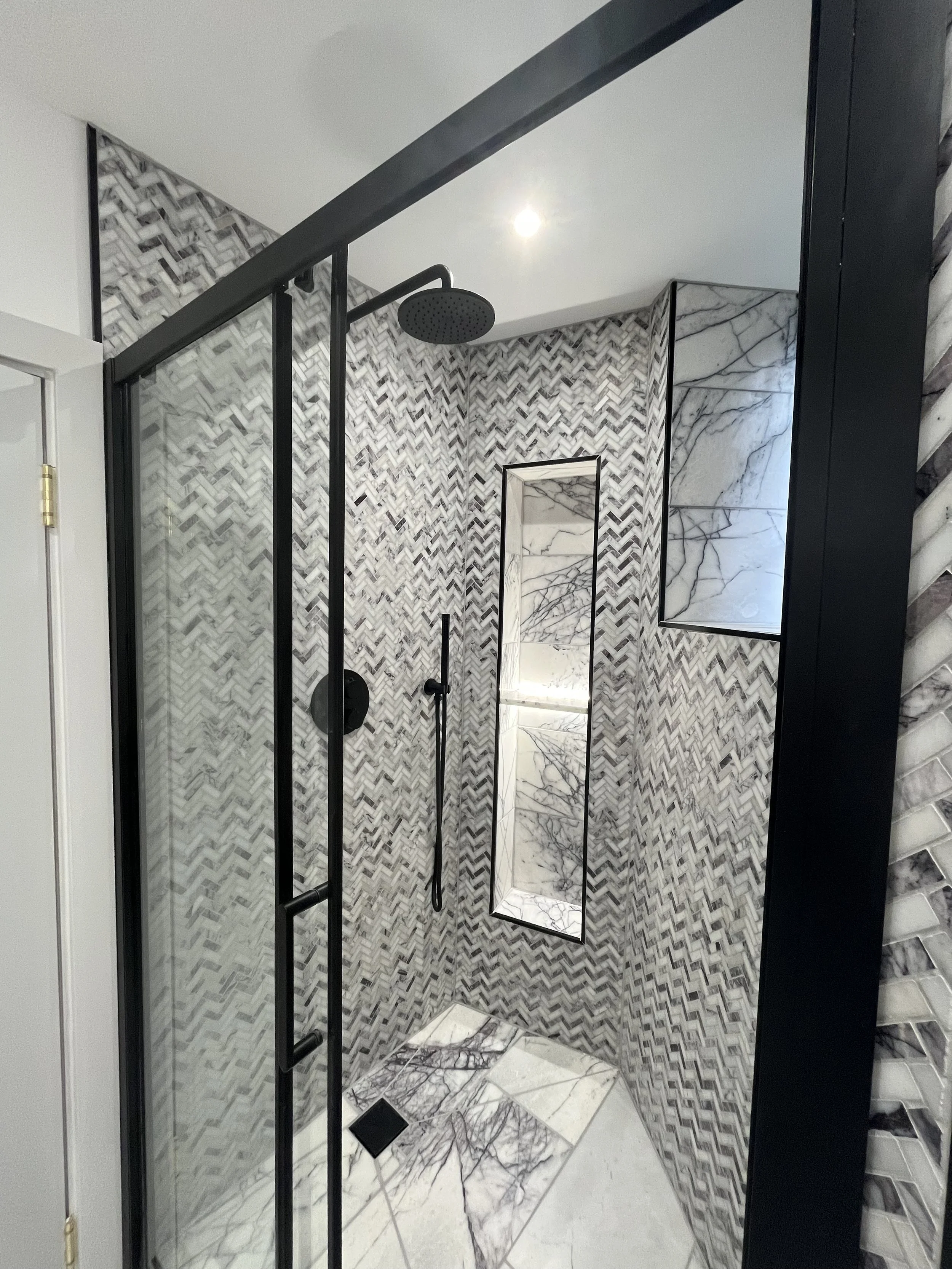 A modern bathroom shower area with patterned tiled walls, a circular rain shower head, a handheld shower head, and a rectangular mirror reflecting marble tiles.
