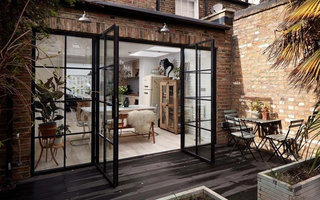 View of a modern indoor-outdoor living space with dark wooden flooring, brick walls, and black-framed glass doors opening to a small outdoor patio with seating, potted plants, and palm trees.