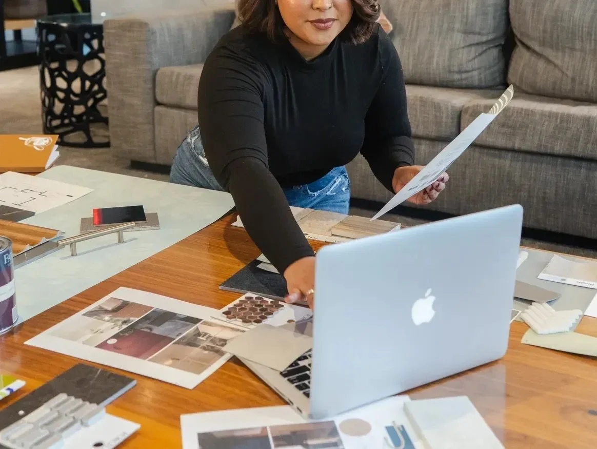 Woman organizing design materials and documents on a wooden table with a silver MacBook, papers, fabric swatches, and color samples, in a modern living room.