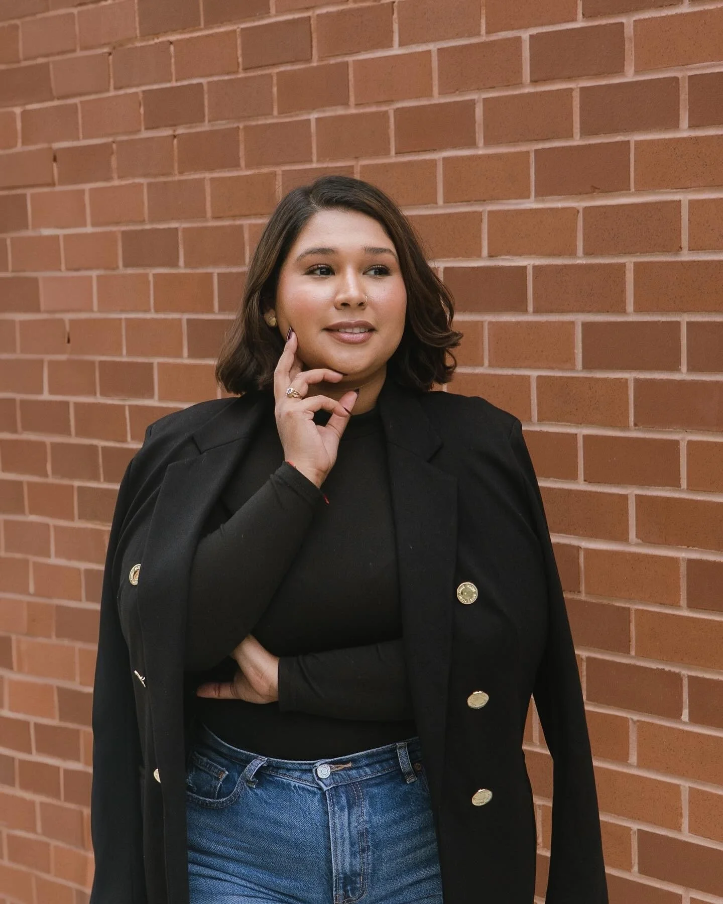 A woman with shoulder-length dark hair and a nose piercing stands in front of a brick wall, wearing a black blazer and jeans, with her hand near her chin, looking to the side.