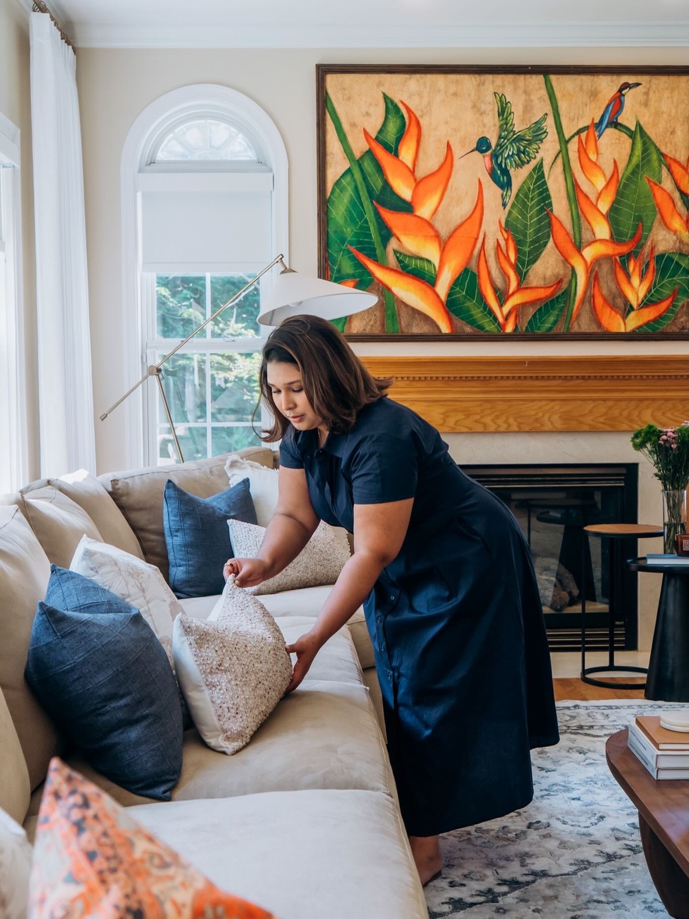 A woman in a dark dress is arranging pillows on a beige couch in a living room with a large floral painting and a window in the background.