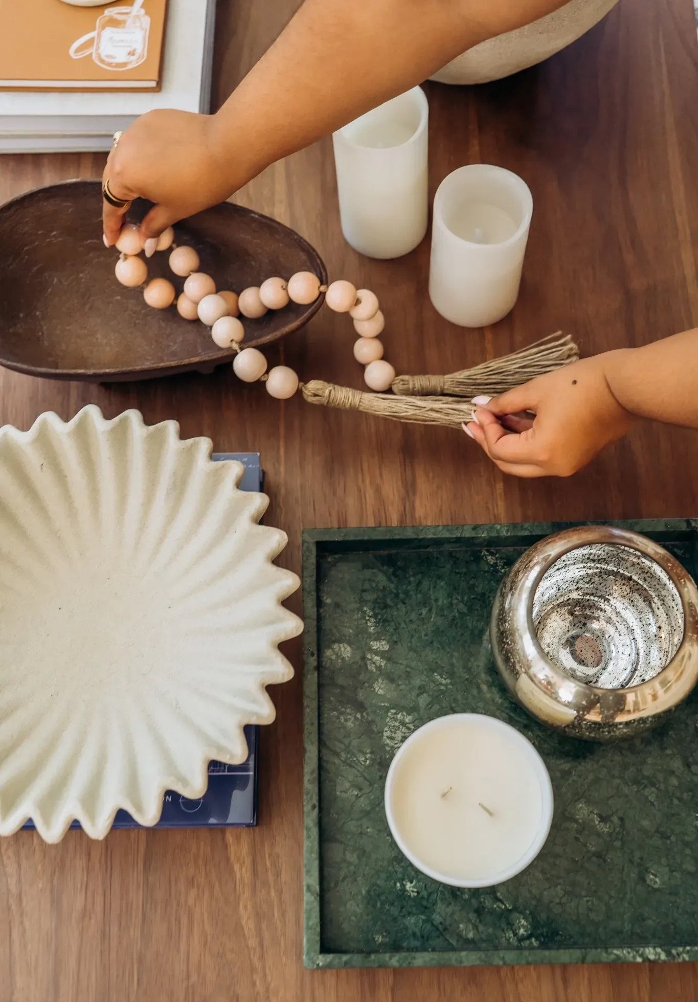 Person arranging a wooden bead necklace on a table with decorative items, two white candles, and ceramic bowls around.
