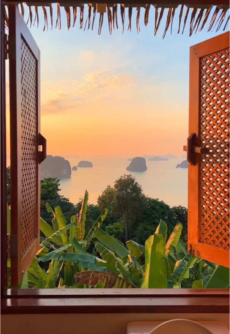 Sunset view through an open window with wooden shutters, tropical greenery in the foreground, and distant islands across calm water.
