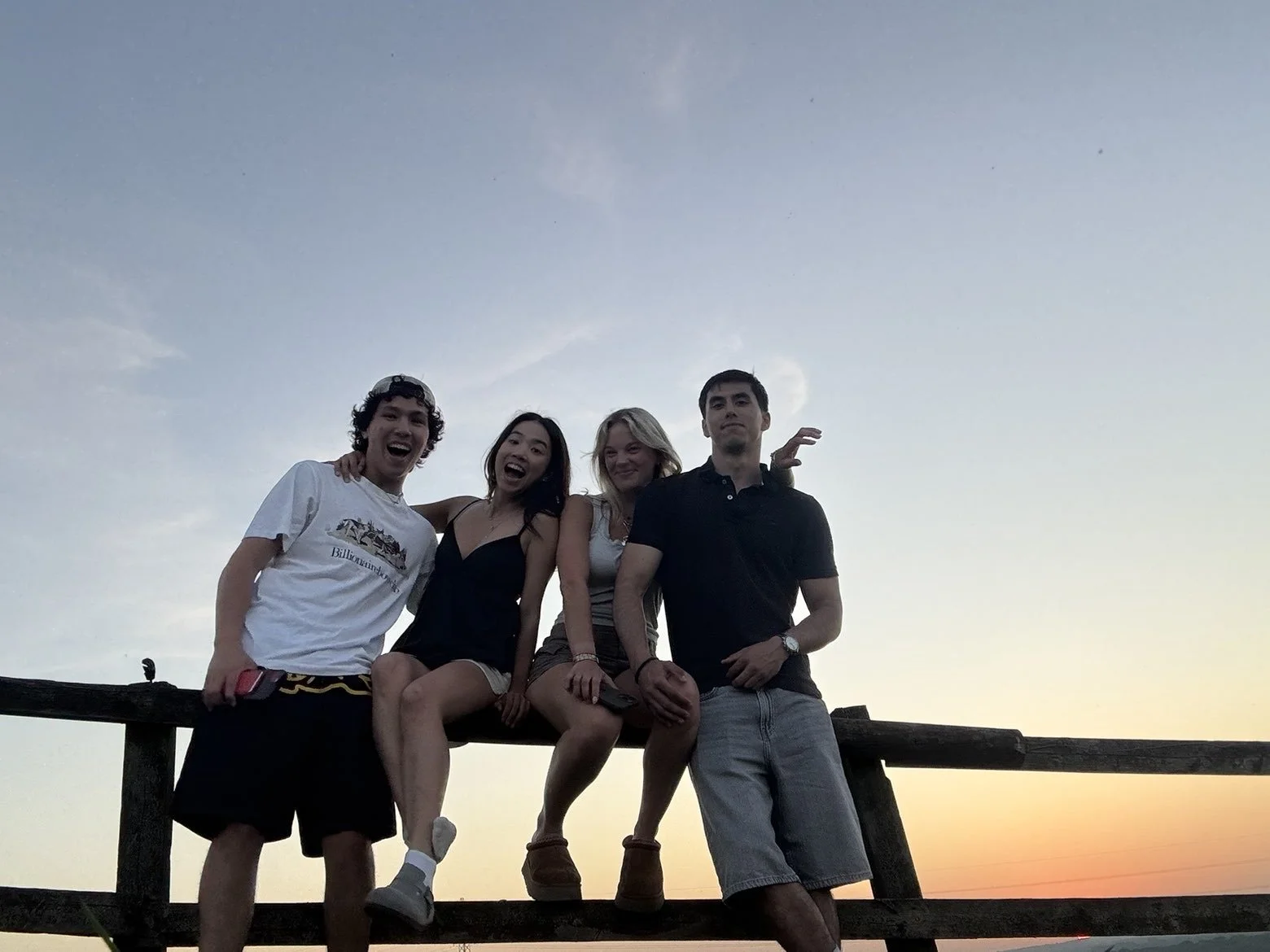 Four friends sitting on a wooden railing outdoors during sunset, smiling and having fun.