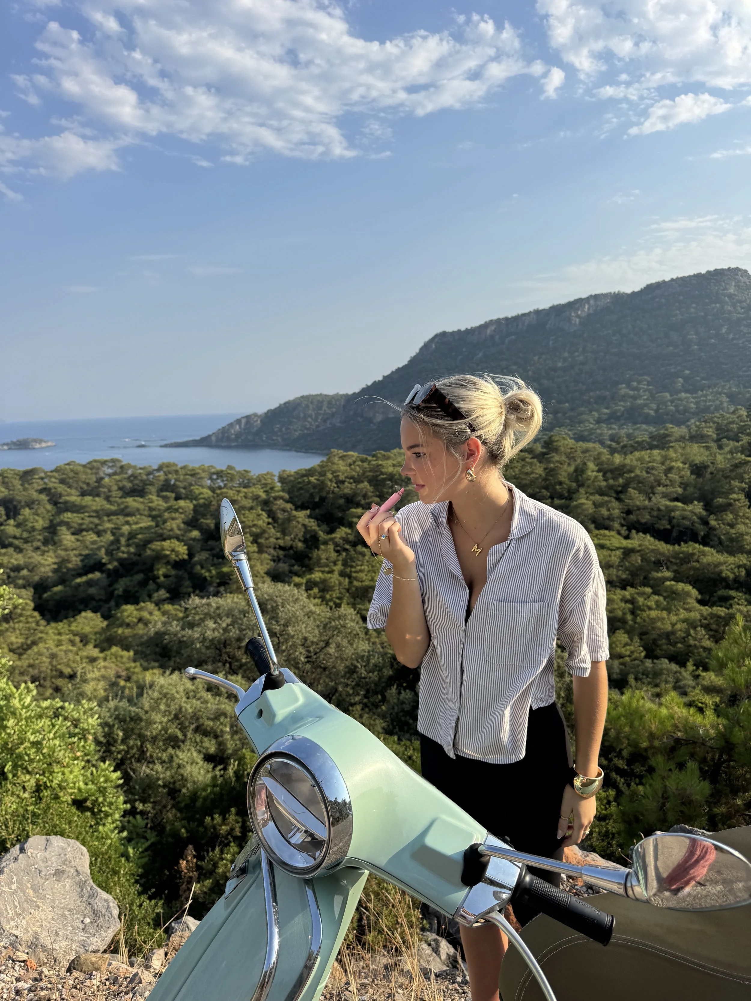 A woman with blonde hair tied back, wearing a striped shirt, sunglasses on her head, and jewelry, stands outdoors near a mint green scooter, with a background of green hills, water, and a partly cloudy sky.