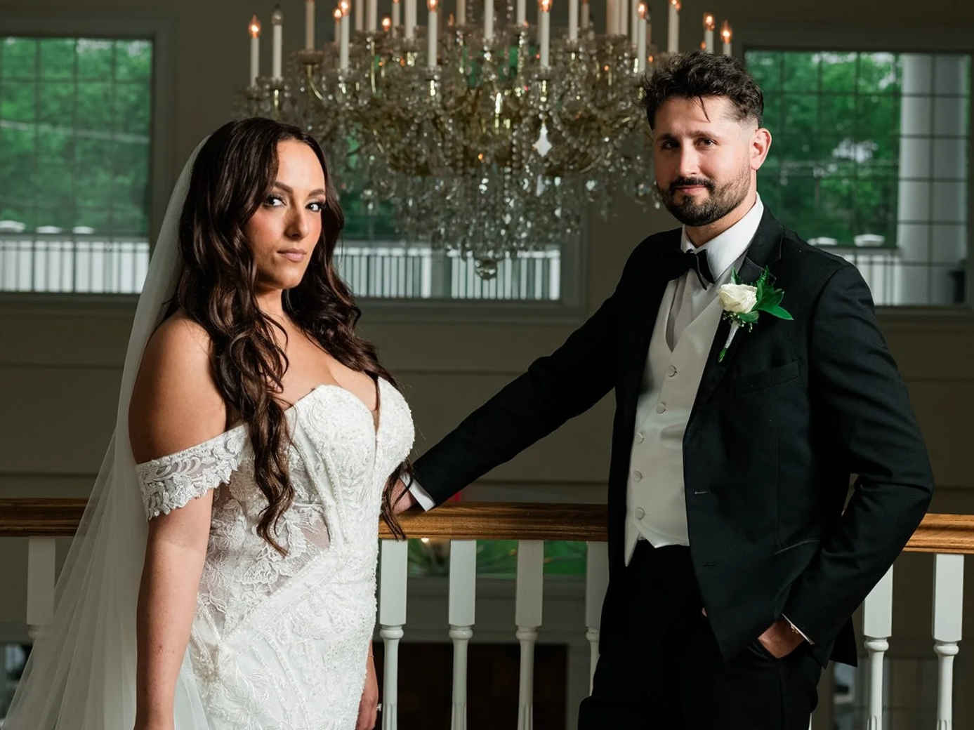 Bride and groom standing indoors in front of a chandelier and windows, dressed in wedding attire, with the bride in a white lace wedding dress and the groom in a black tuxedo with a white shirt and boutonniere.