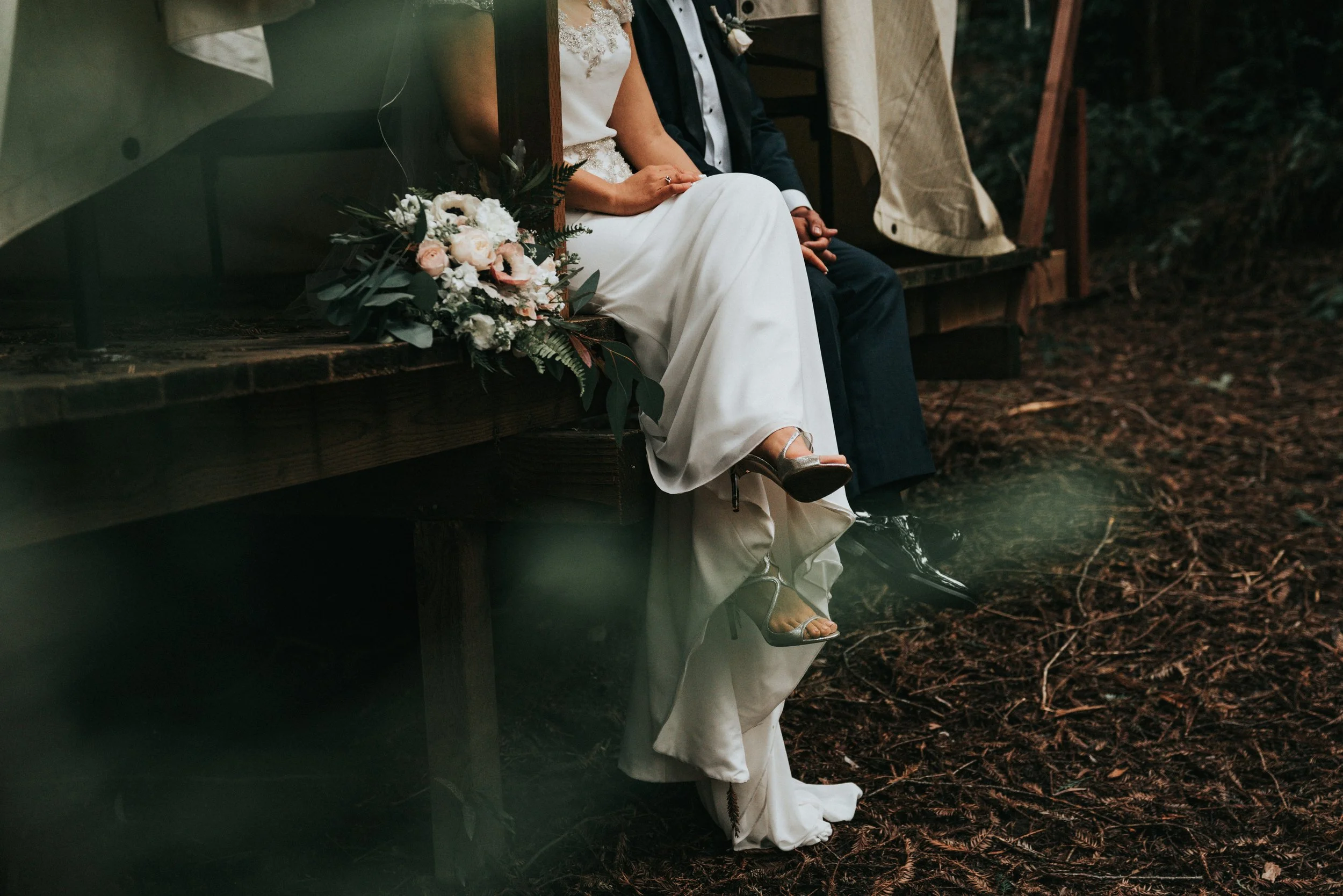 A newlywed couple sitting on a wooden bench outdoors, with a bouquet of flowers beside them, partially visible, during a wedding ceremony.