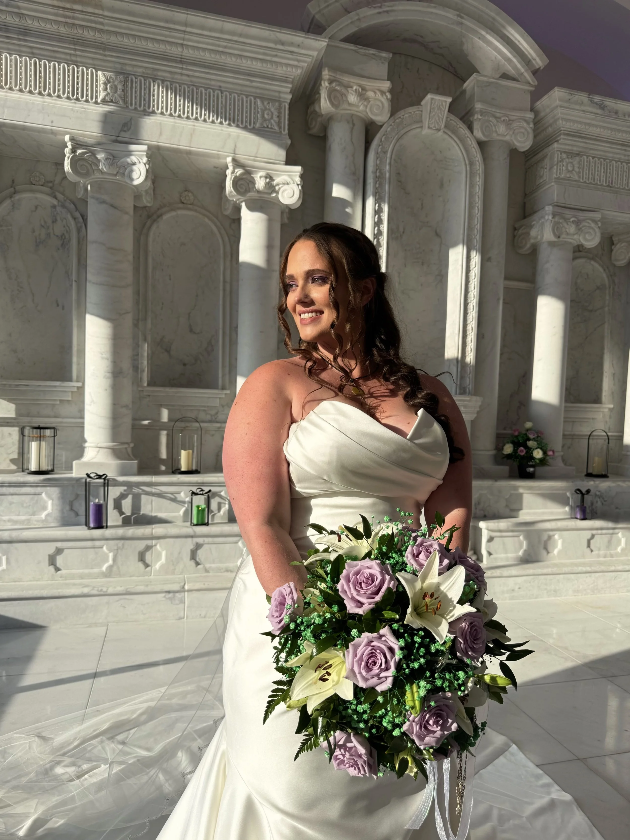A woman in a white wedding dress holding a bouquet of lavender roses, white lilies, and greenery, standing inside a grand marble hall with columns and decorative architectural features.
