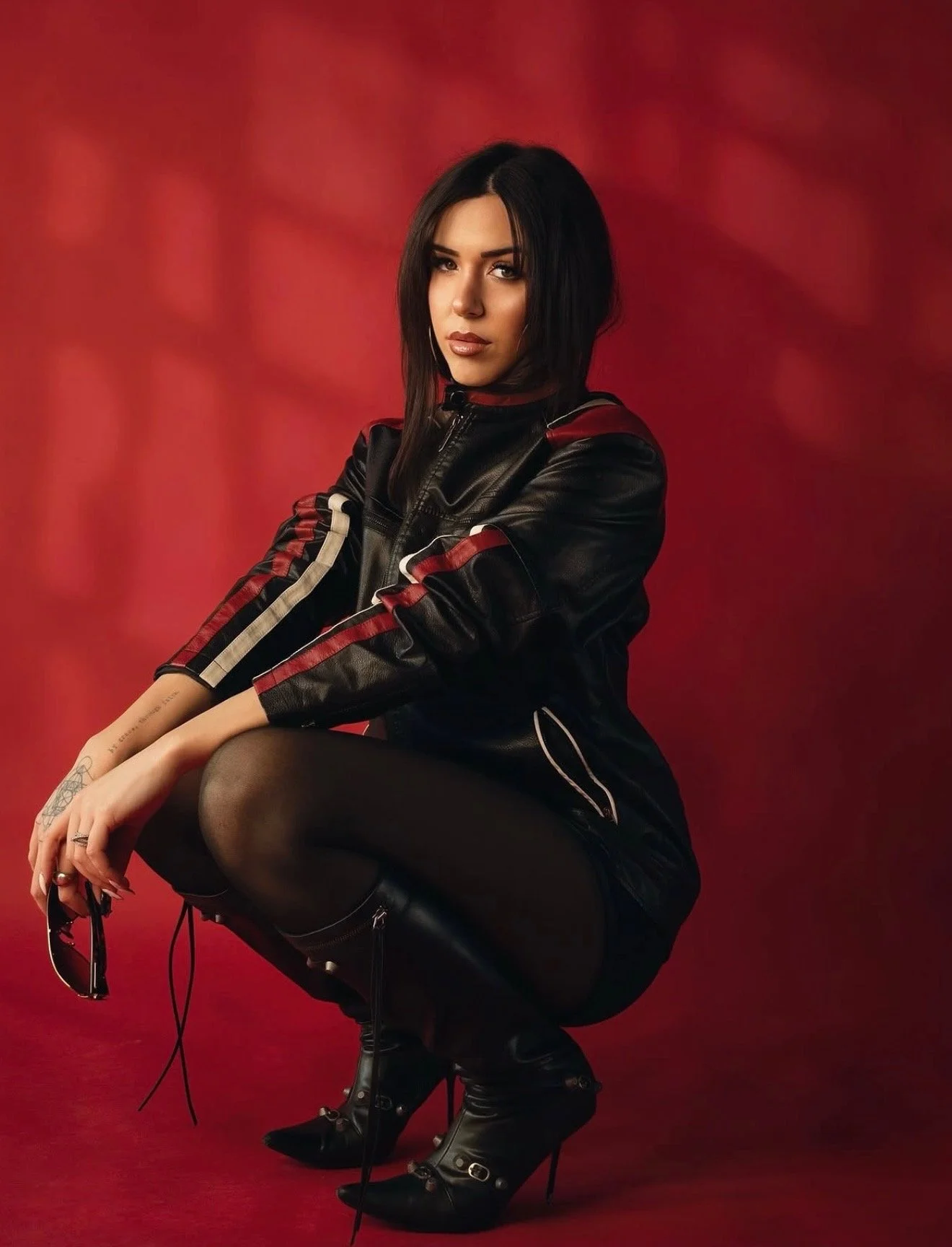 A woman with black hair and makeup, wearing a black leather jacket with red and white stripes, black skirt, and high-heeled black boots, sitting against a red background.