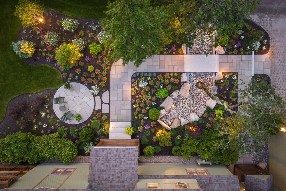 Aerial view of a landscaped garden with flower beds, trees, a stone pathway, and a sitting area with chairs.